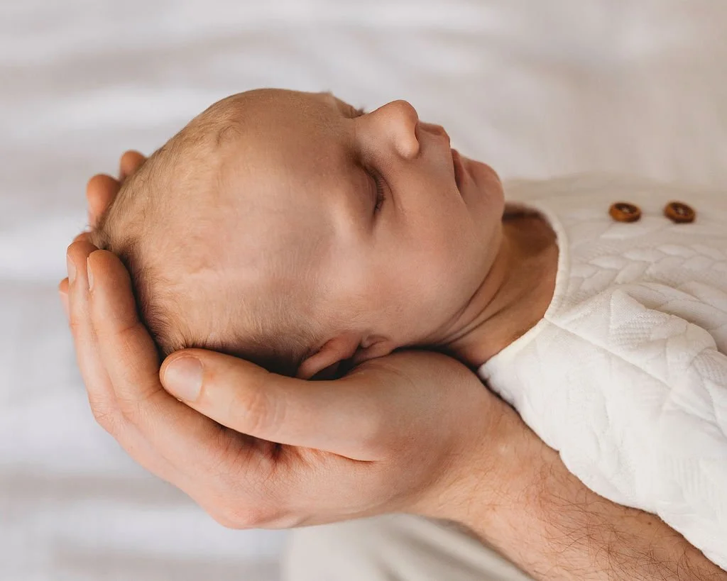 A baby with closed eyes, resting peacefully in an adult's hand and arm, with a white textured blanket or clothing.