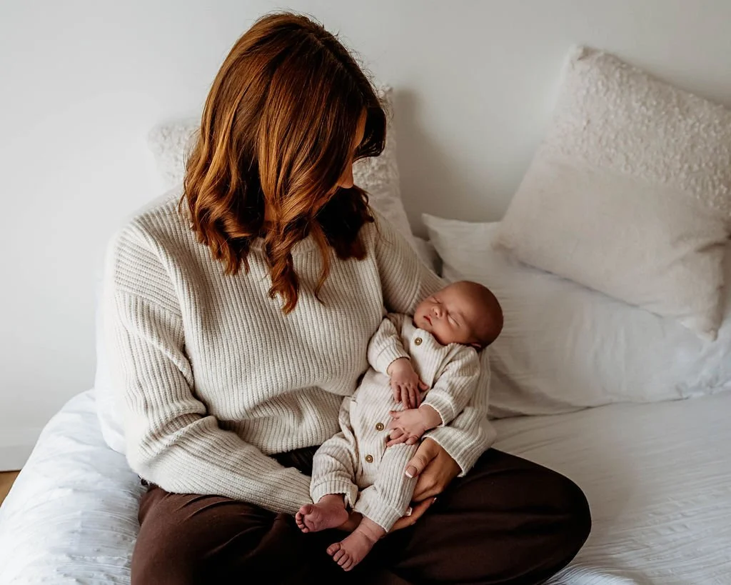 A woman sitting on a bed holding a sleeping baby in her arms.