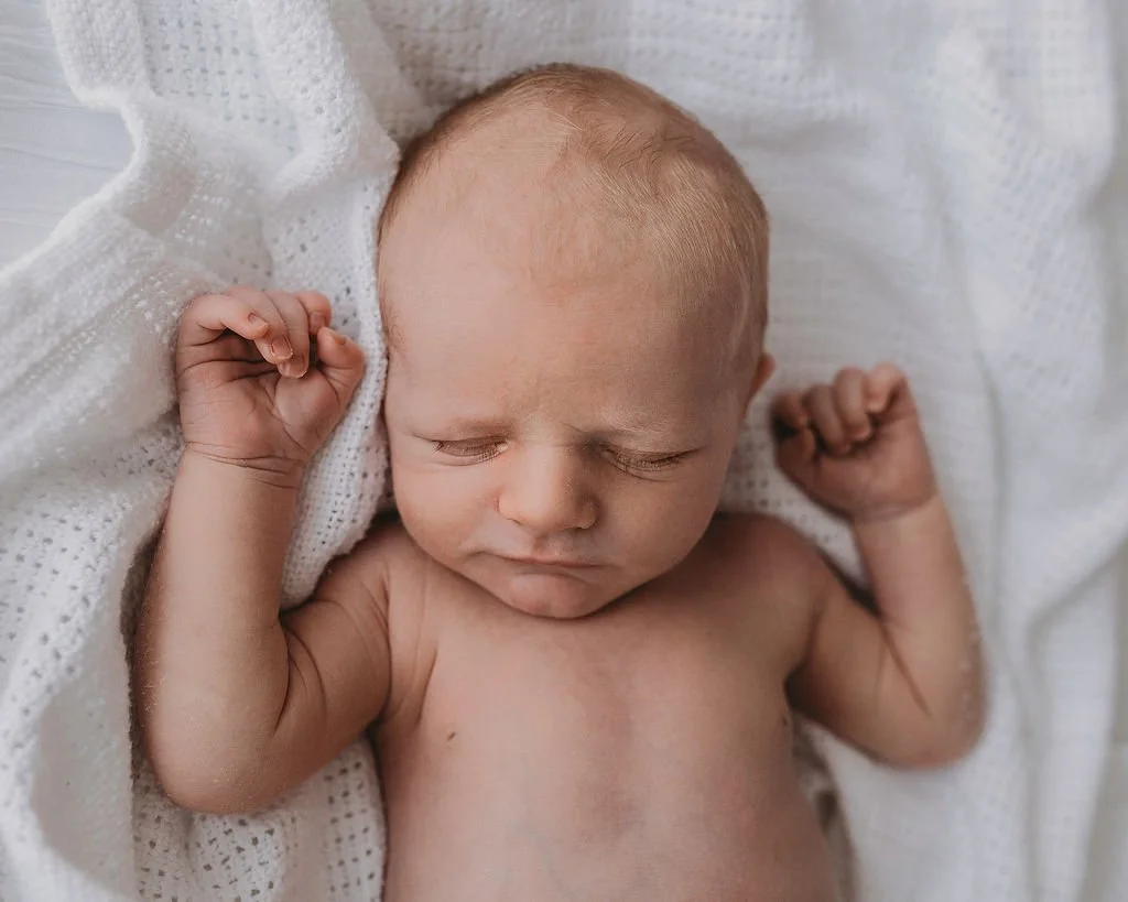 A sleeping baby with closed eyes, on a white blanket, with arms raised and fists clenched.