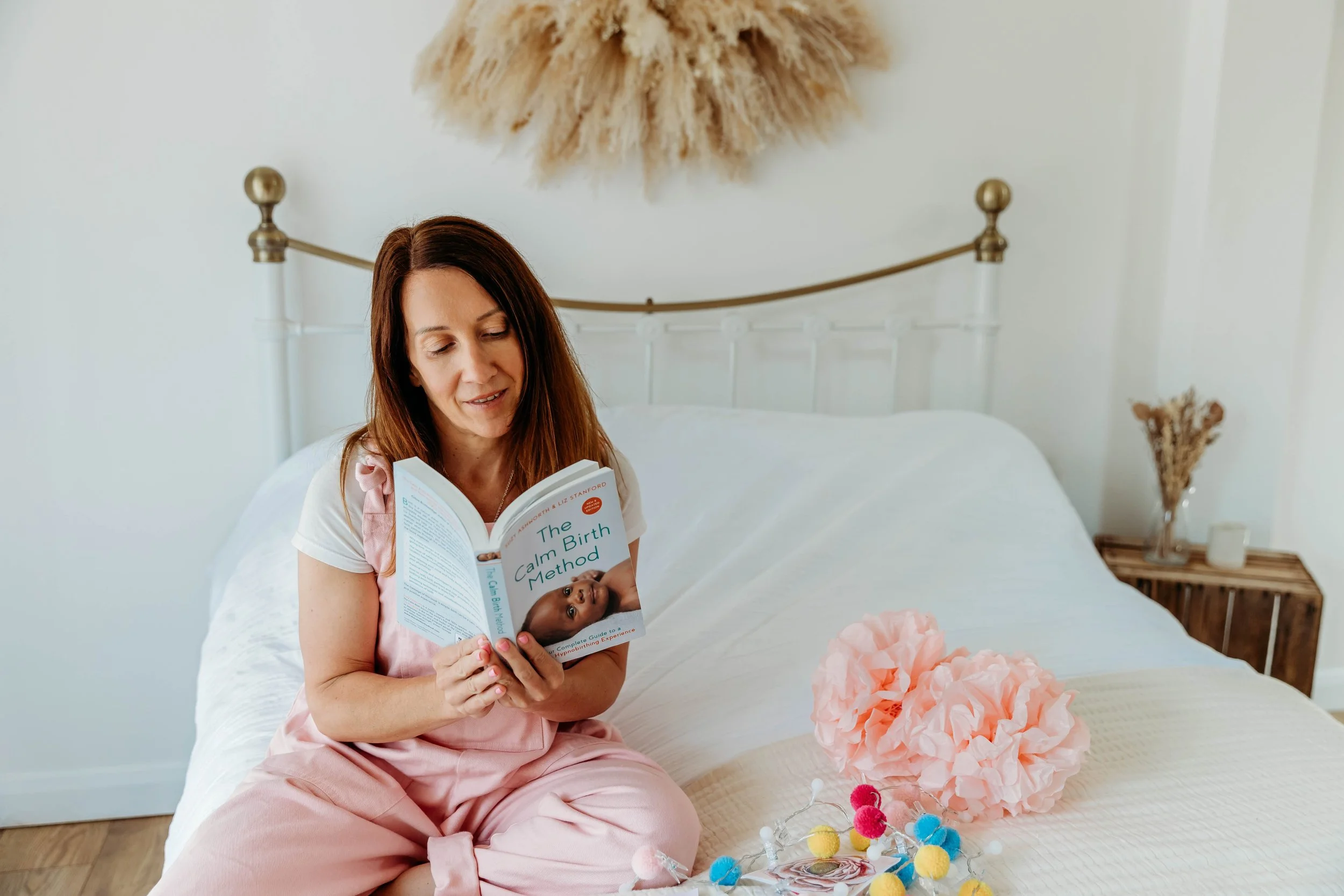 A woman sitting on a bed, reading a book titled 'The Calm Birth Method' in a cozy bedroom with pink and pastel decor.