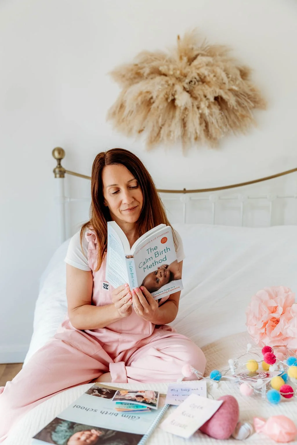 Woman in pink pajamas reading 'The Calm Birth Method' book while sitting on a white bed with baby photos, pink pom-pom lights, and soft toys around her in a cozy bedroom.