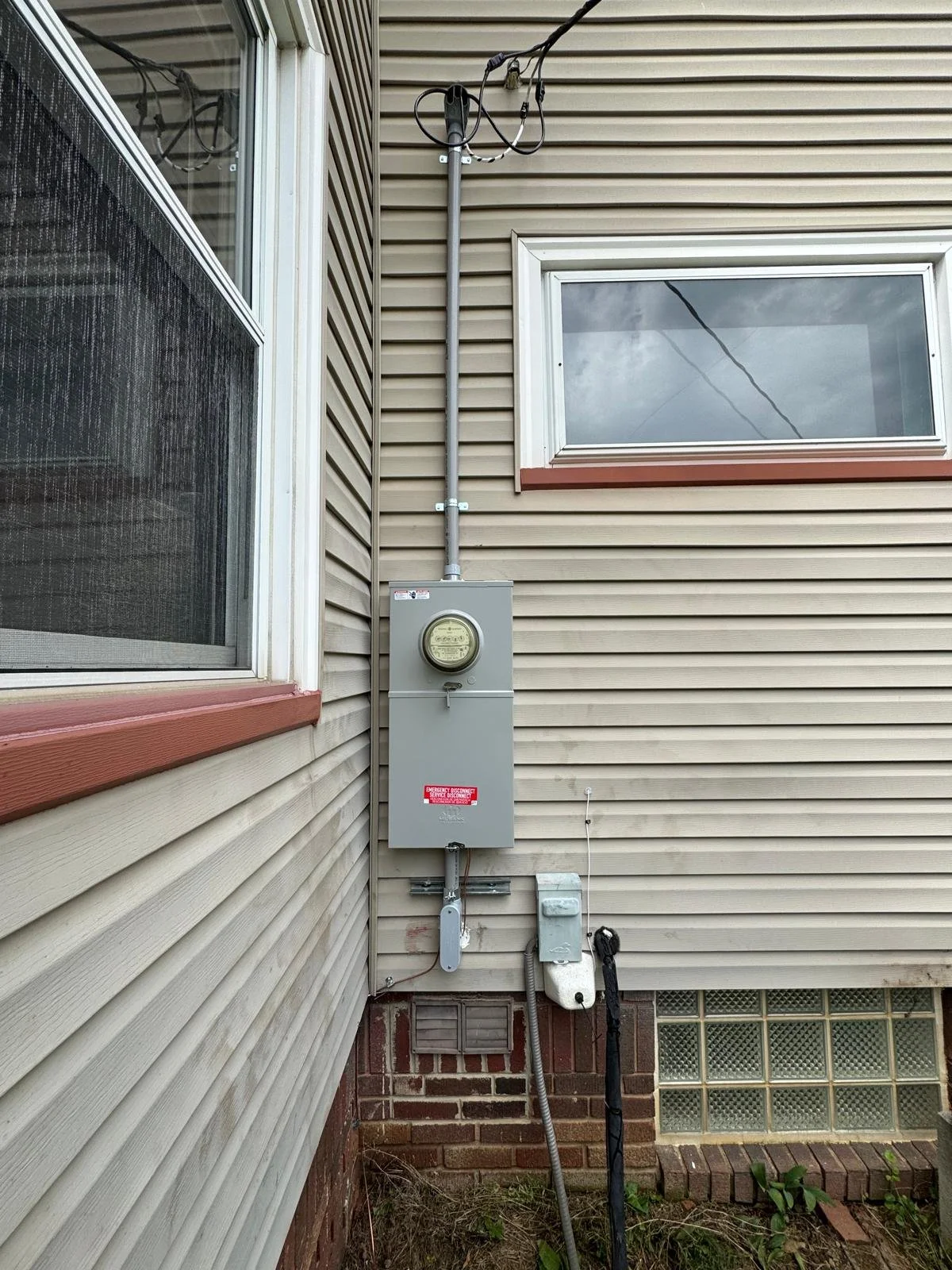 Electrical meter and box mounted on the exterior wall of a house, with power lines overhead and windows nearby.