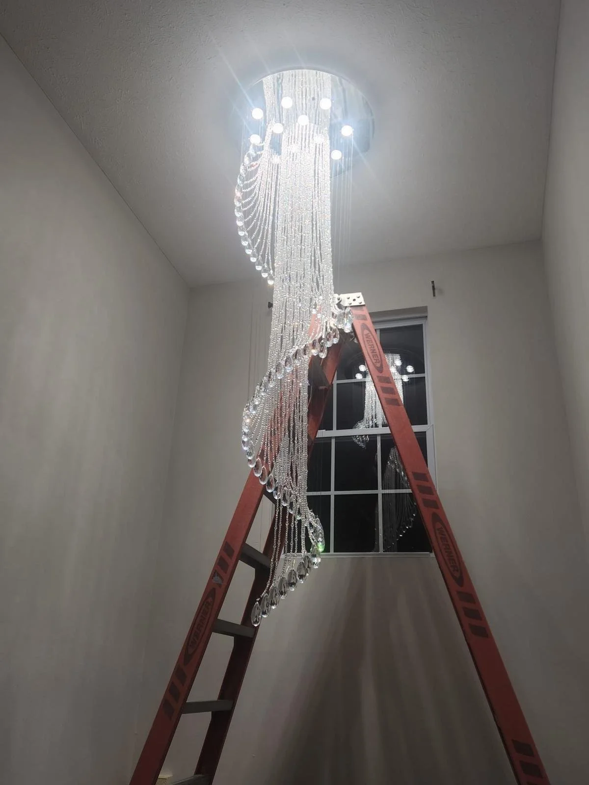 A sparkling chandelier hanging from the ceiling, illuminated by a light source at the top, with a ladder leaning against the wall in front of a window.