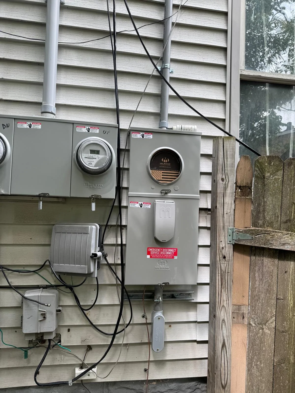 Utility meters and electrical boxes mounted on the exterior of a house wall, next to a wooden fence.