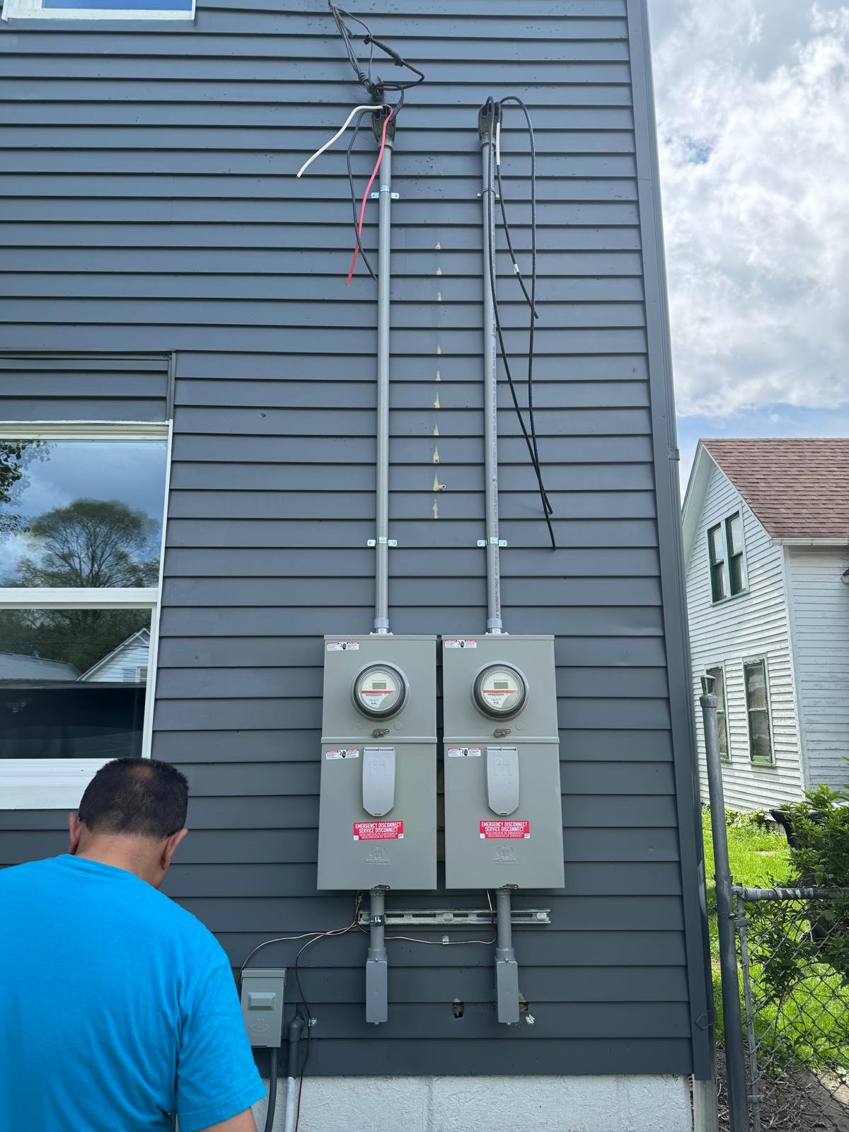 Installation of electrical meters with wiring on the exterior wall of a house, a man in a blue shirt working near the meters.