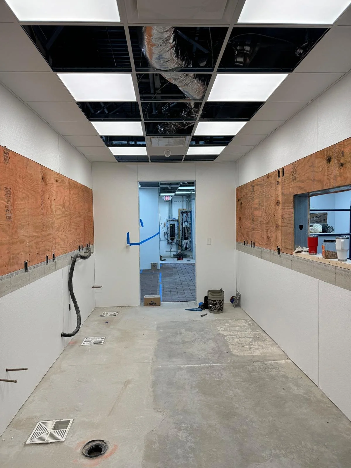 A room under construction with exposed ceiling tiles, some missing, showing ductwork. The walls have plywood panels and wiring. The floor is unfinished concrete with vents and a drain. There are tools, buckets, and construction materials scattered on