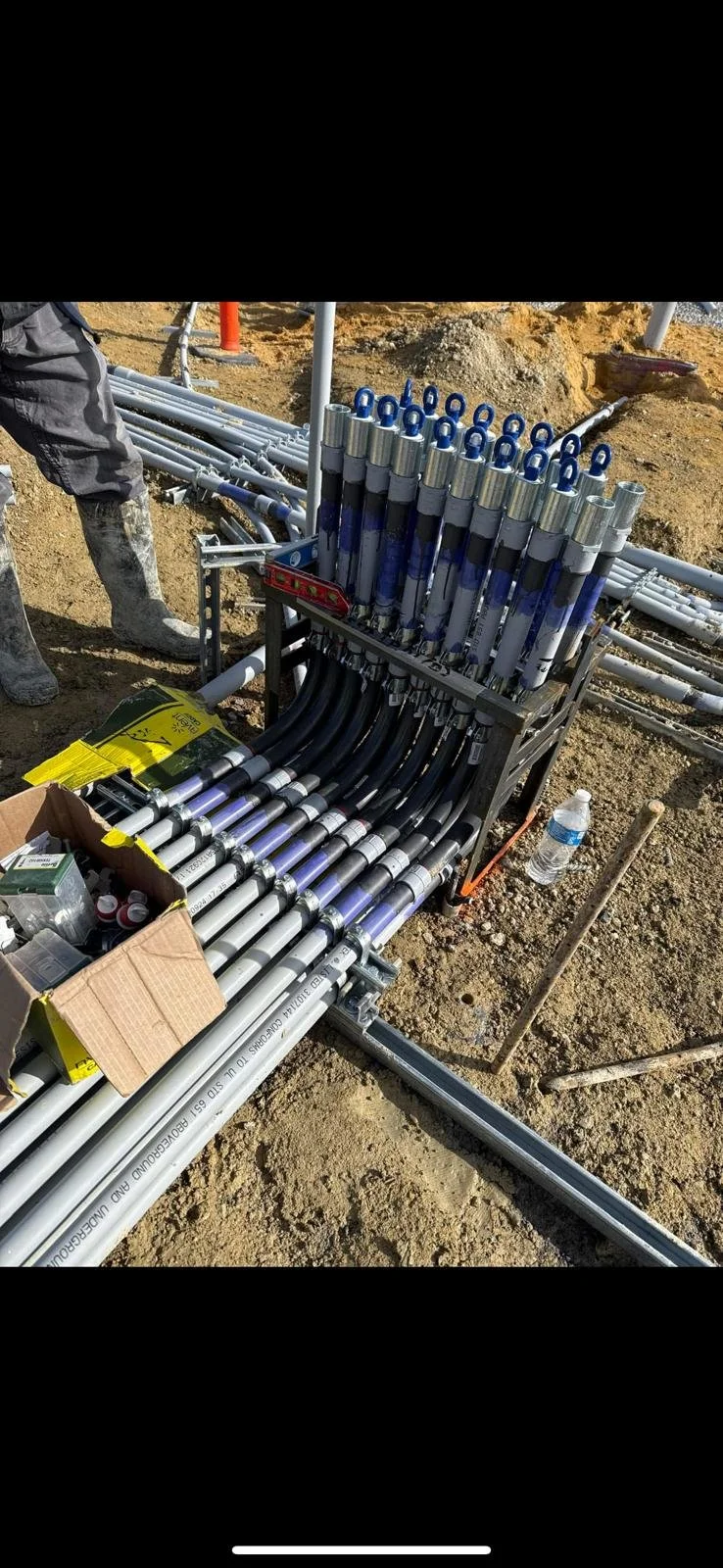 Construction site with pipes and tubing organized in a cart and on the ground, with a person’s legs partially visible, a box of supplies, and a water bottle.