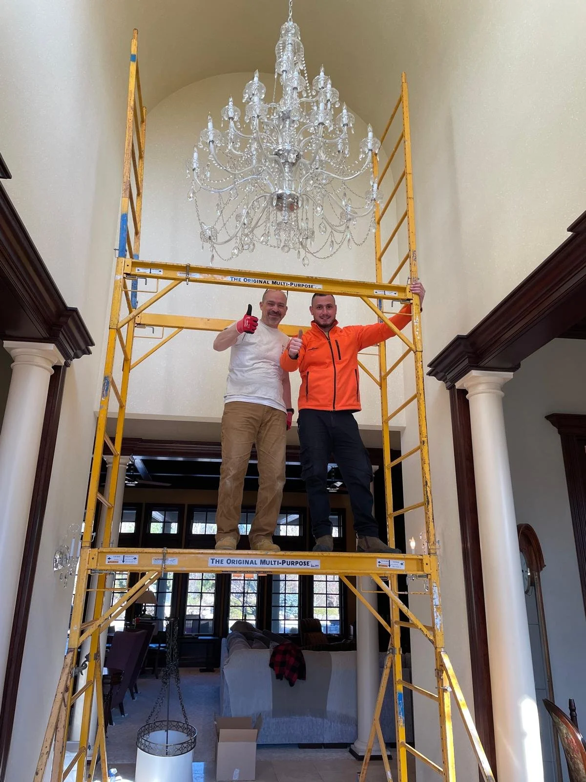 Two men standing on a yellow scaffold inside a spacious house, giving thumbs up. One man wears a white shirt, the other an orange jacket. A large crystal chandelier hangs above them.