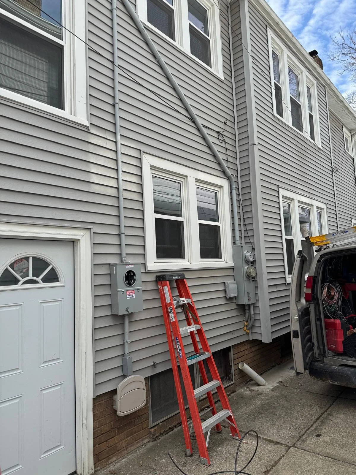 Exterior of a multi-story house with gray vinyl siding, multiple windows, and electrical meters. A red ladder is leaning against the house, and a utility van with open doors is parked nearby.