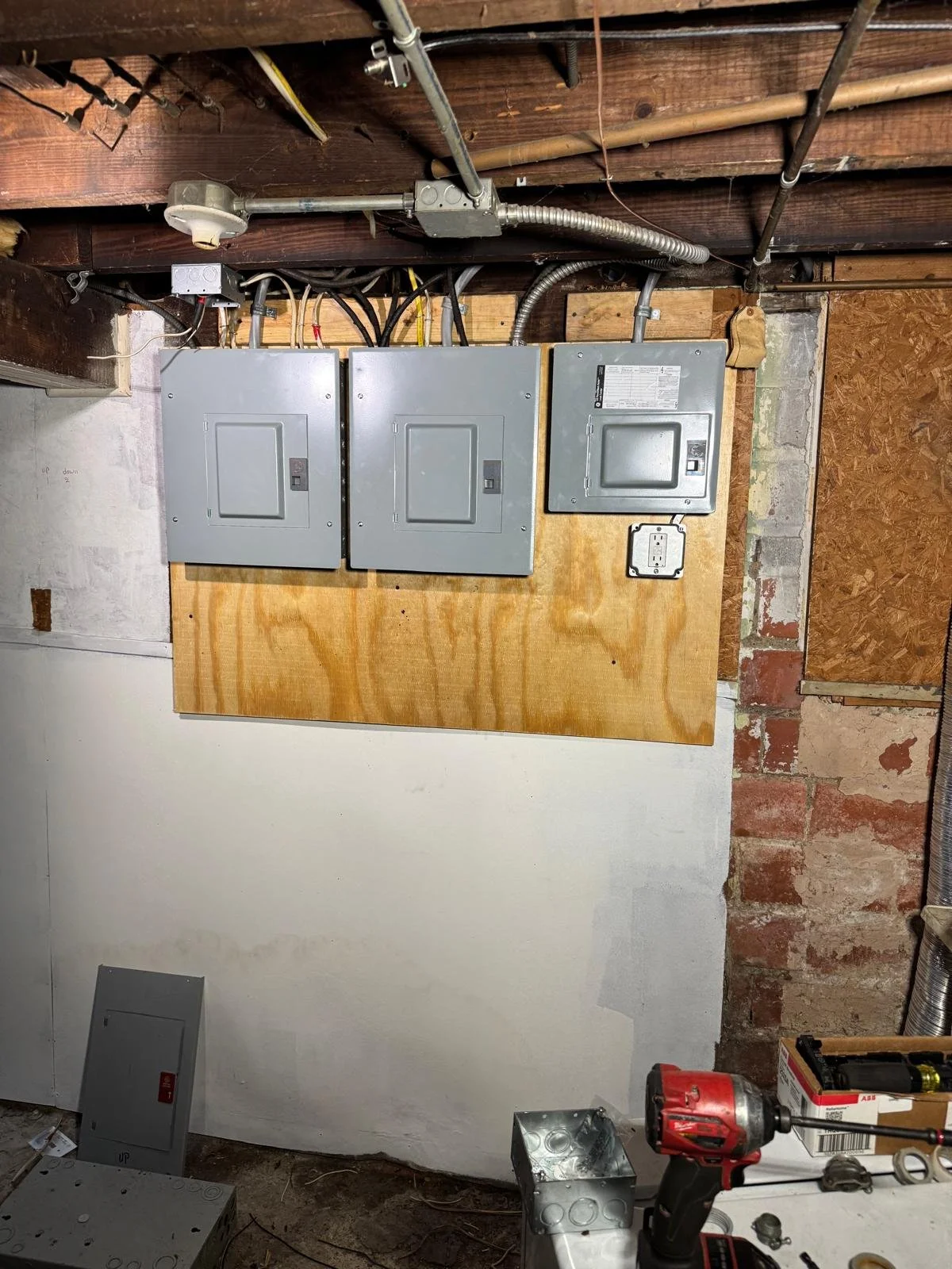 Electrical panel with three gray breaker boxes mounted on a plywood wall, with wiring and conduit above, in a garage or workshop setting.