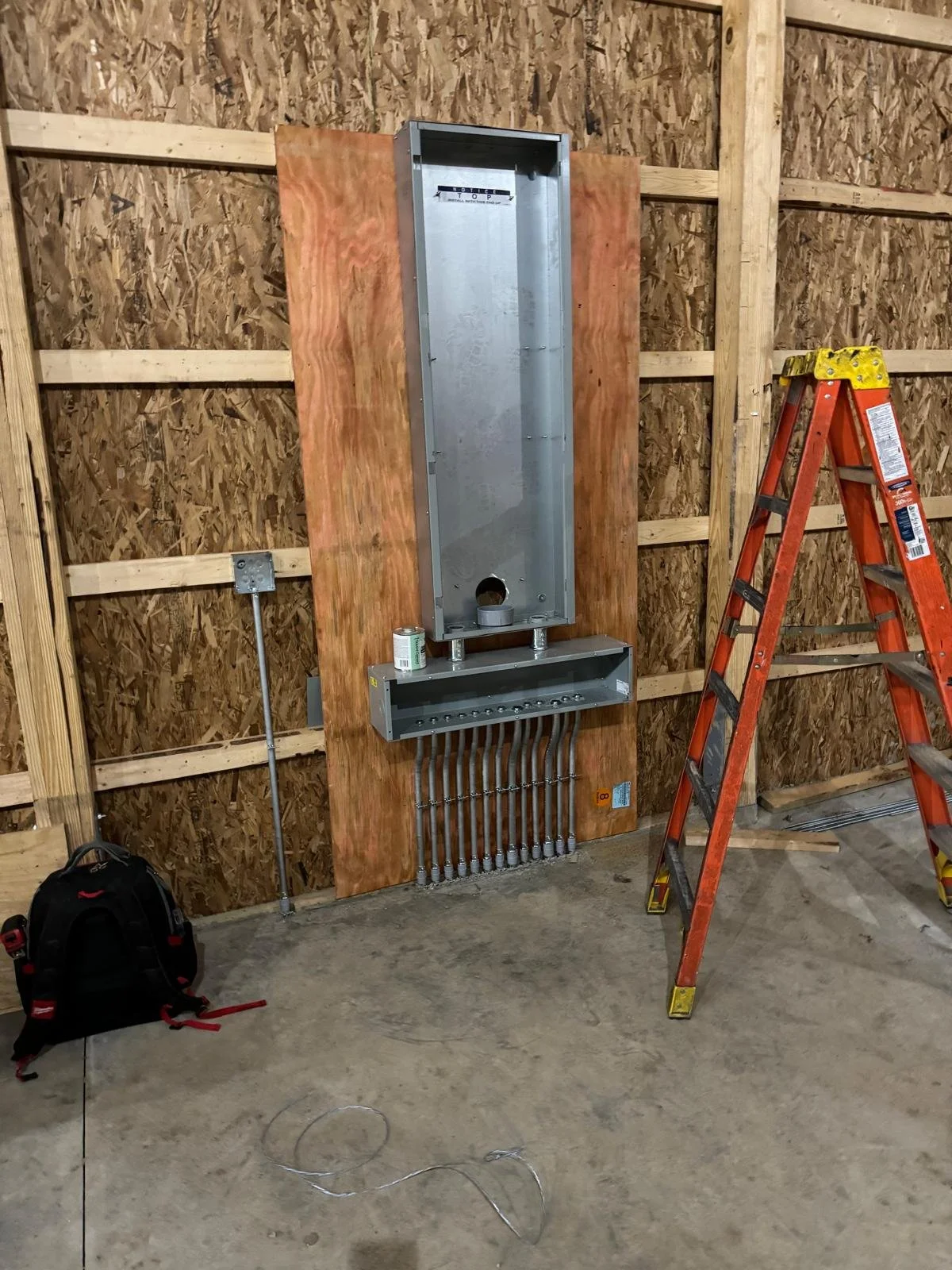 Electrical wiring box installation in a partially finished building, with an orange ladder, a backpack, and a can of paint or lubricant nearby.