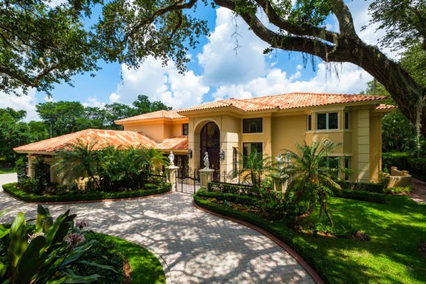Luxury two-story house with a tiled roof, surrounded by lush greenery and tall trees on a sunny day.