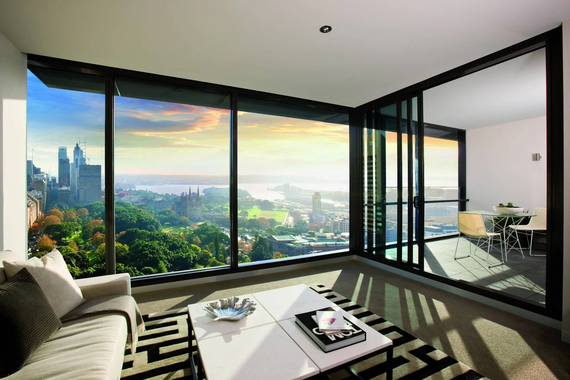 Modern living room with large floor-to-ceiling windows offering a city skyline and park view, with a seating area and a small balcony with a table and chairs.