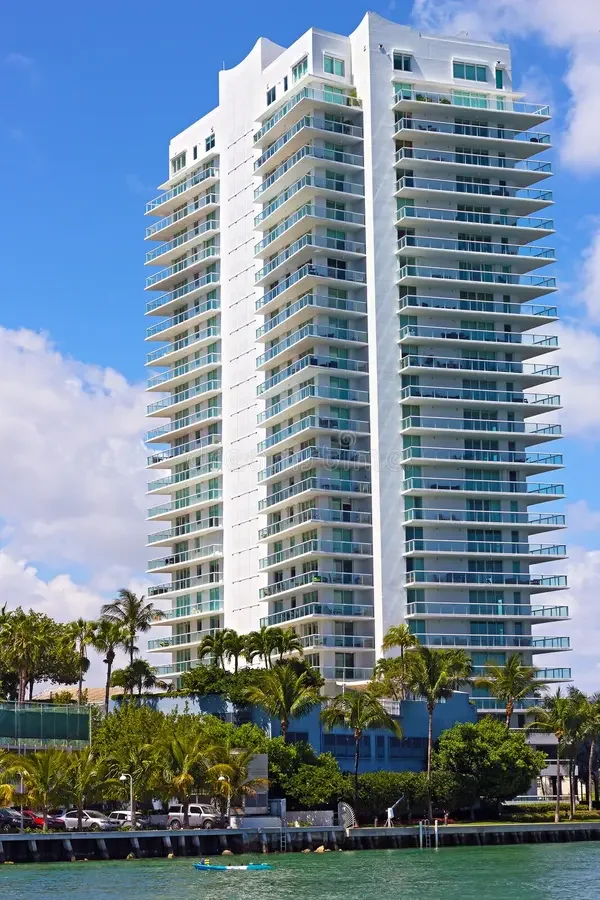A tall modern white residential building with multiple balconies, set against a blue sky with some clouds. There are palm trees at the base, along with a waterfront and parked cars visible in the foreground.