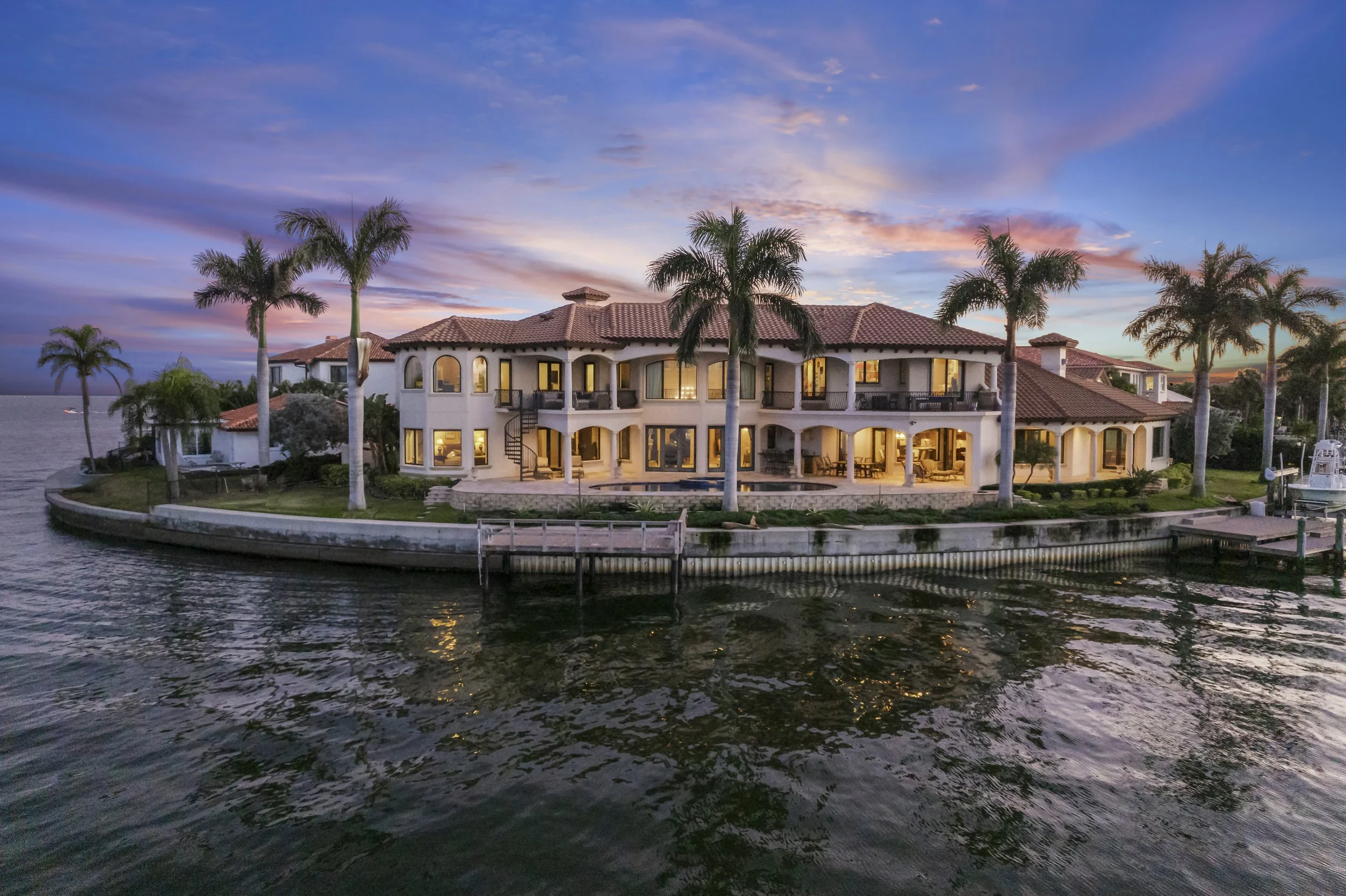 Luxury waterfront house with multiple floors, large windows, surrounded by palm trees, during sunset, with a boat dock in the water.