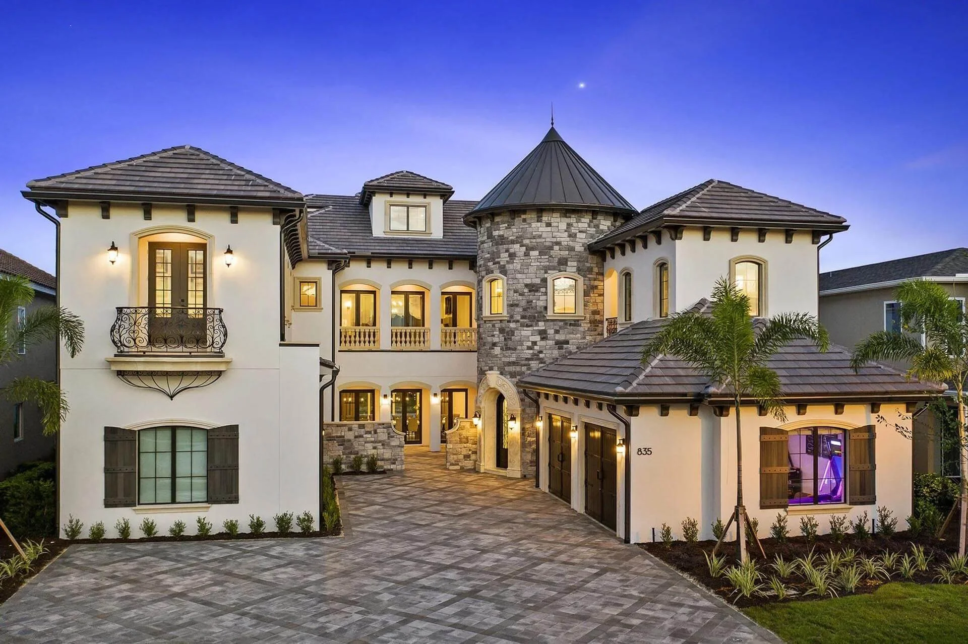 Large elegant house with stone and stucco exterior, multiple windows, and a drive leading to a three-car garage, illuminated in evening light with palm trees and landscaping.