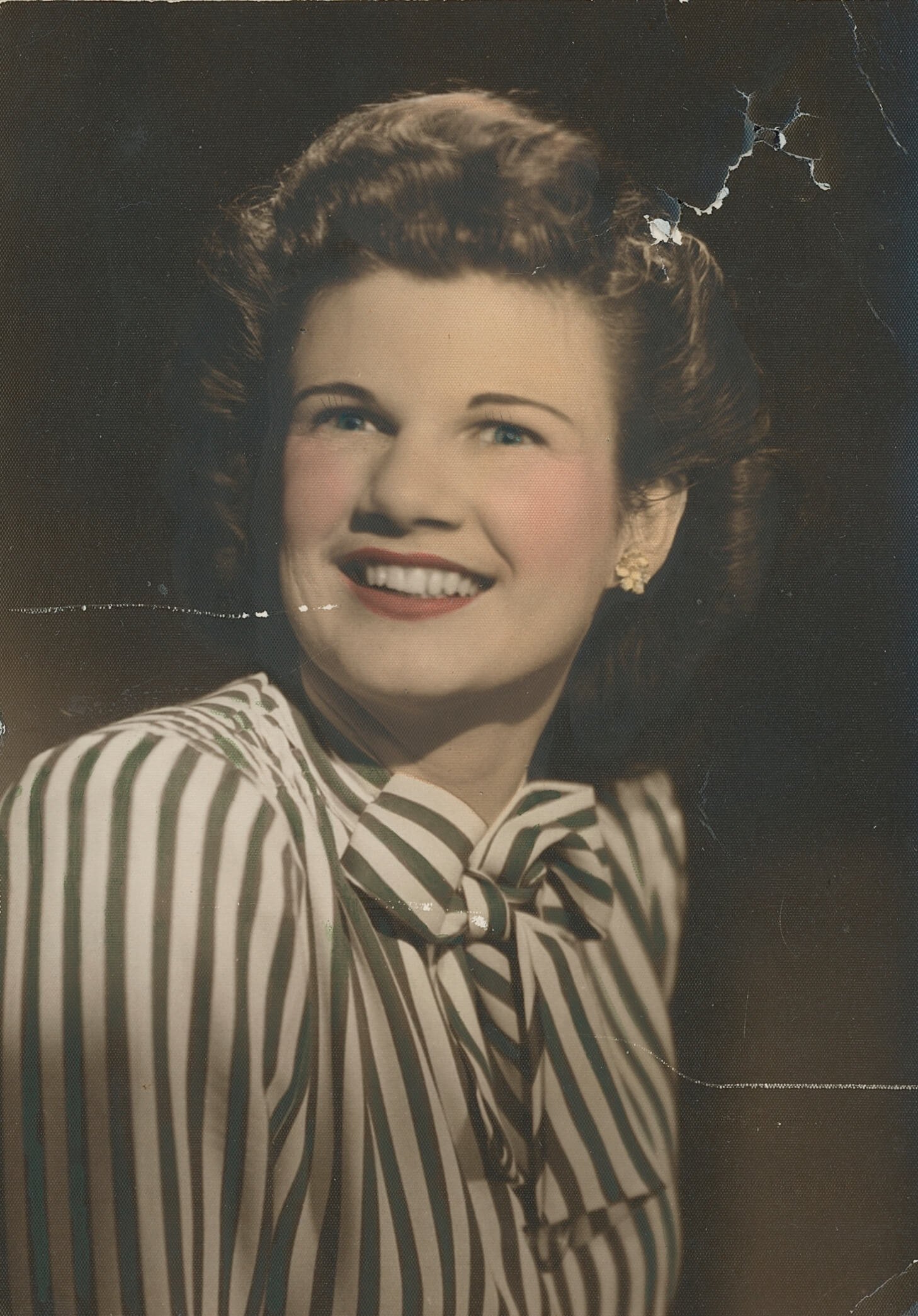 Old colorized portrait of a smiling woman in a striped blouse with visible damage, scratches, and fading, representing a photo before restoration.