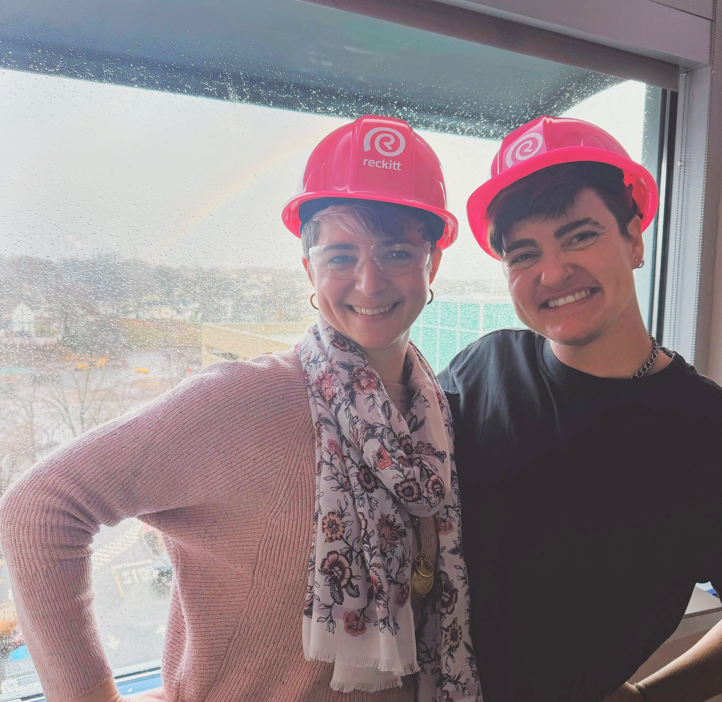 Two smiling individuals wearing pink construction helmets standing in front of a rain-speckled window, with a cityscape background visible through the window.