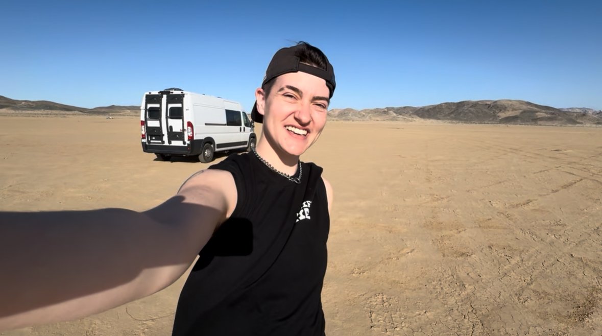 Young person taking selfie in a desert with a white van in the background and distant hills, wearing a black cap backwards, a black sleeveless shirt, and a chain necklace.