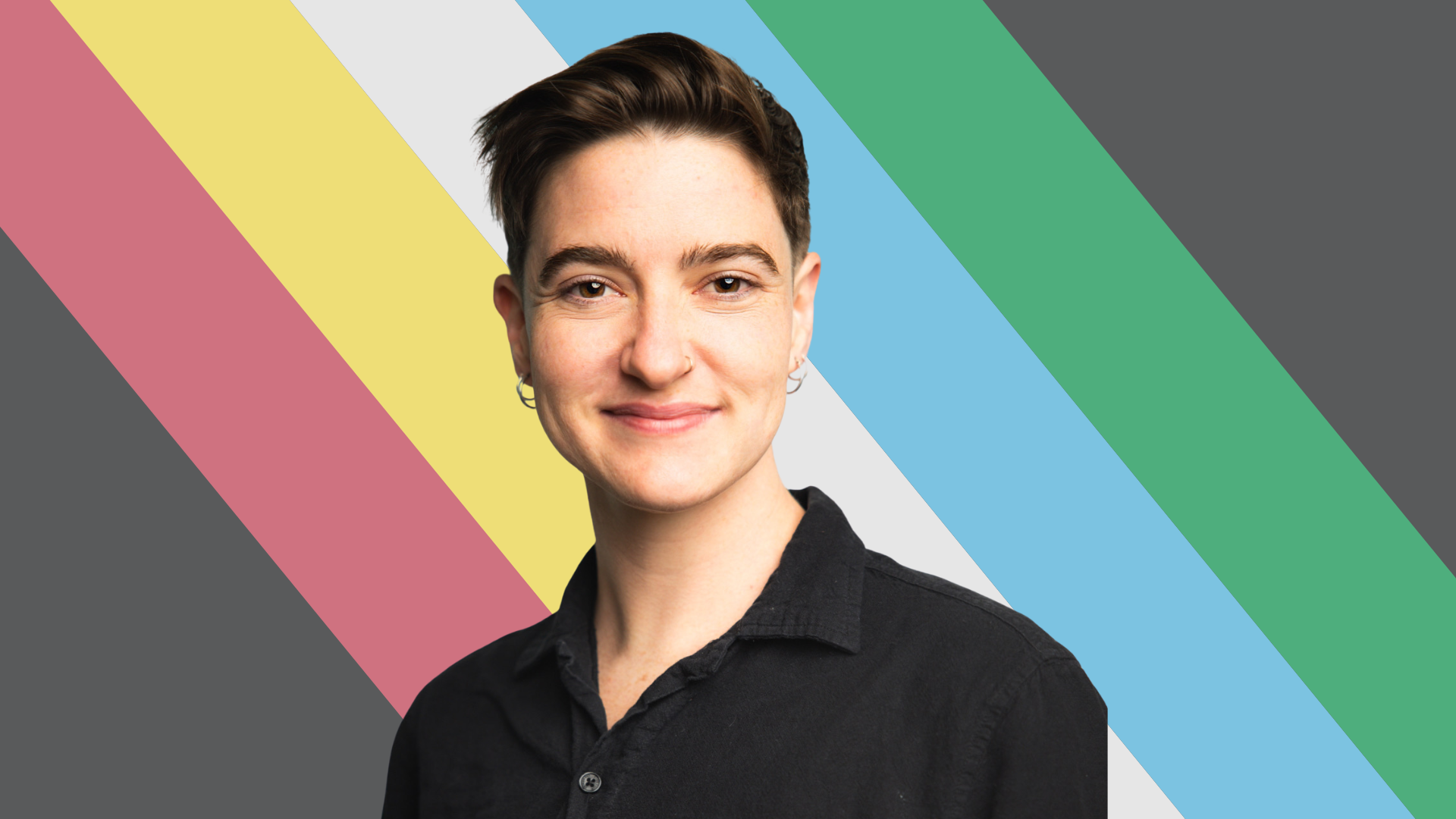 A person with short dark hair smiling, wearing a black shirt, with a background of diagonal rainbow-colored stripes.