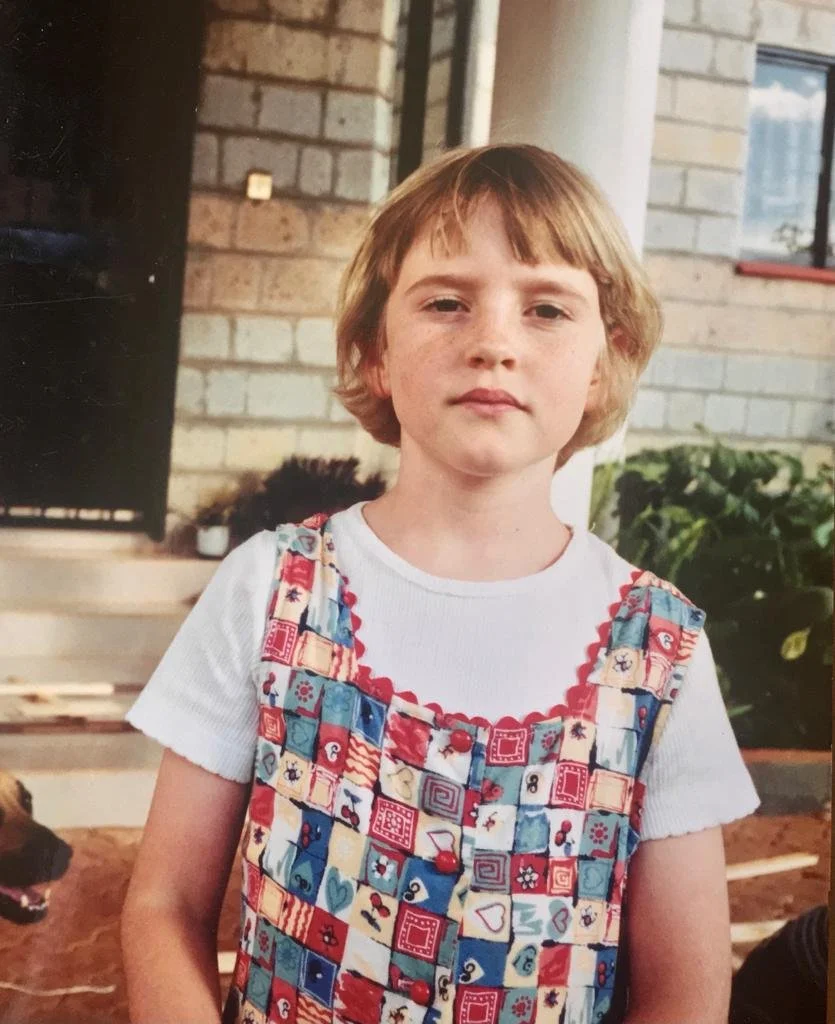 A young girl with short, light brown hair and freckles standing outdoors. She is wearing a white t-shirt and a colorful patchwork dress with red, blue, and cream patterns. There is a brick building and some greenery in the background.