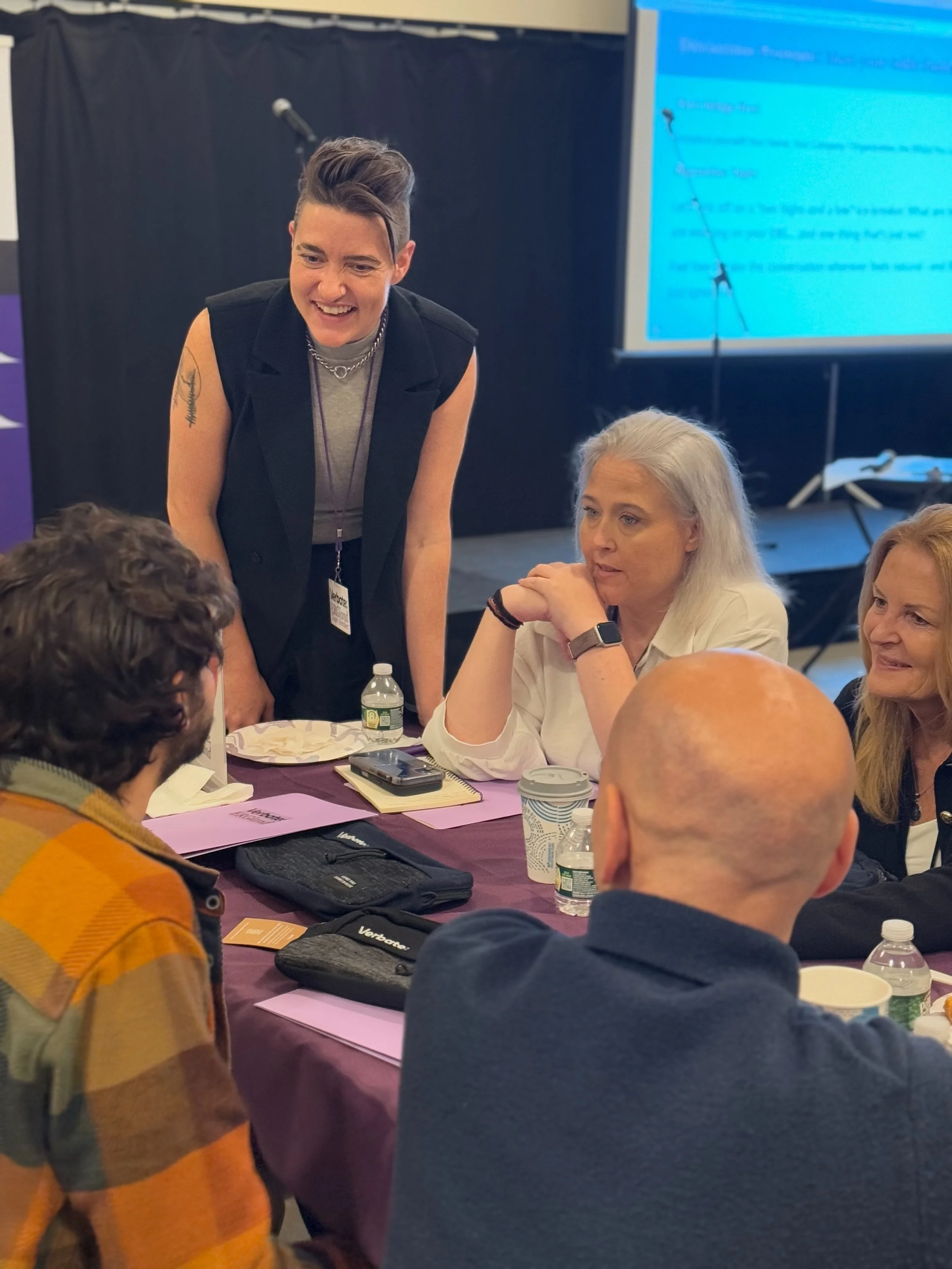 A group of people sitting around a table in a meeting room, smiling and laughing while a woman stands and talks to them, holding a microphone.
