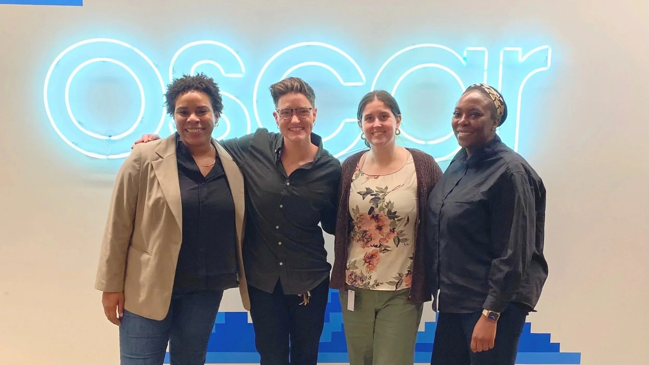 Four women standing together in front of a neon sign that reads 'OASCAR'. They are smiling and have their arms around each other.