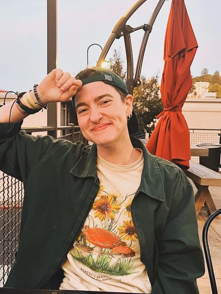 A young person with short dark hair, wearing a black cap backwards, a dark green jacket, and a cream-colored T-shirt with a mushroom and flower print, sitting outdoors at a restaurant or cafe during sunset, smiling and saluting.