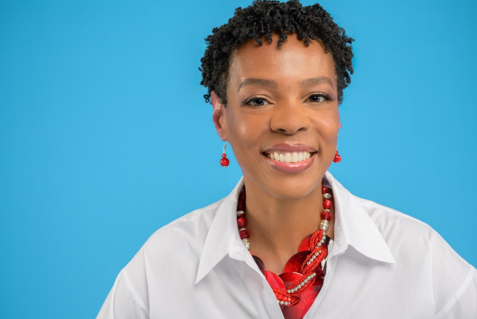 A woman with short curly hair smiling at the camera, wearing a white shirt and red jewelry including earrings, necklace, and bracelet, against a blue background.