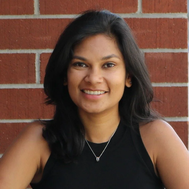 A woman with black hair smiling, wearing a black sleeveless top and a silver necklace, standing in front of a brick wall.