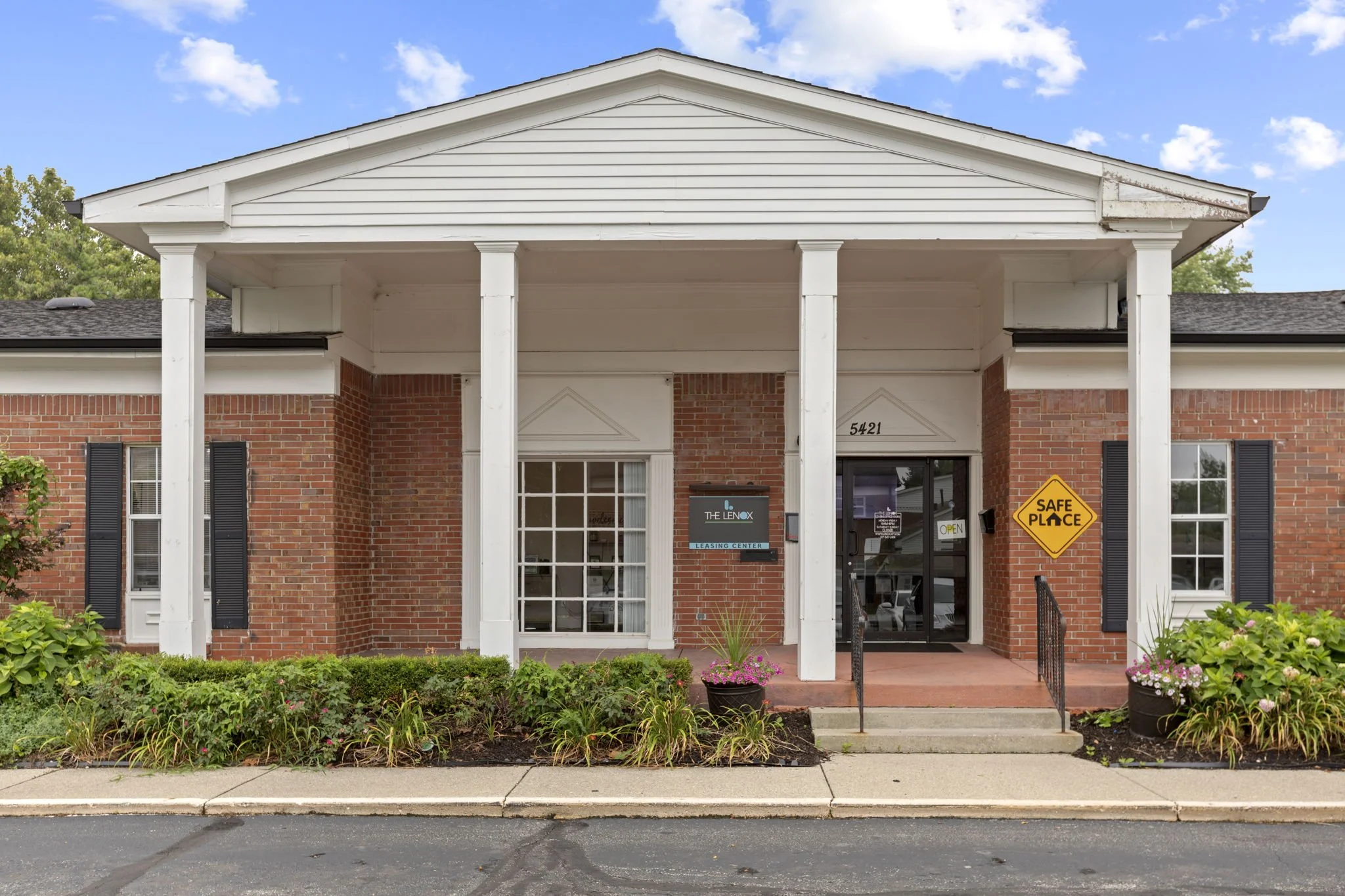 Exterior view of a brick office building with white columns and black shutters, displaying signs that read 'The Lenox Leasing Center' and a yellow warning sign that says 'Safe Place', with a small garden and sidewalk in front.