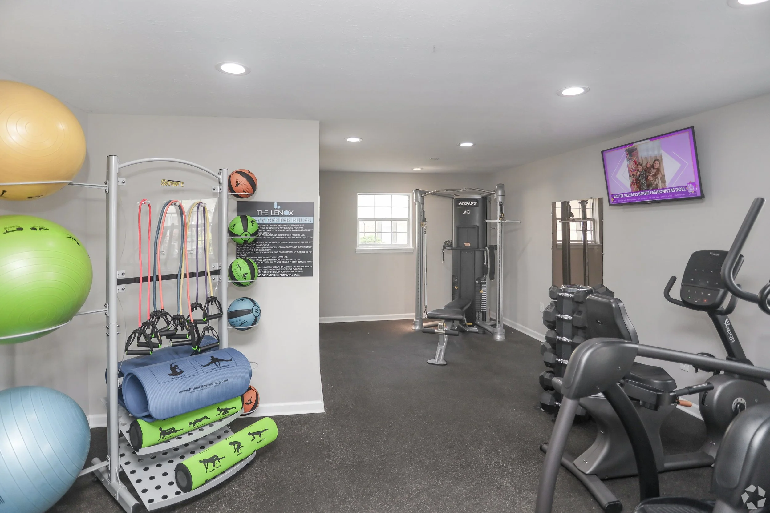 Home gym room with exercise equipment including stability balls, resistance bands, workout mats, weight machine, exercise bike, and a wall-mounted TV.