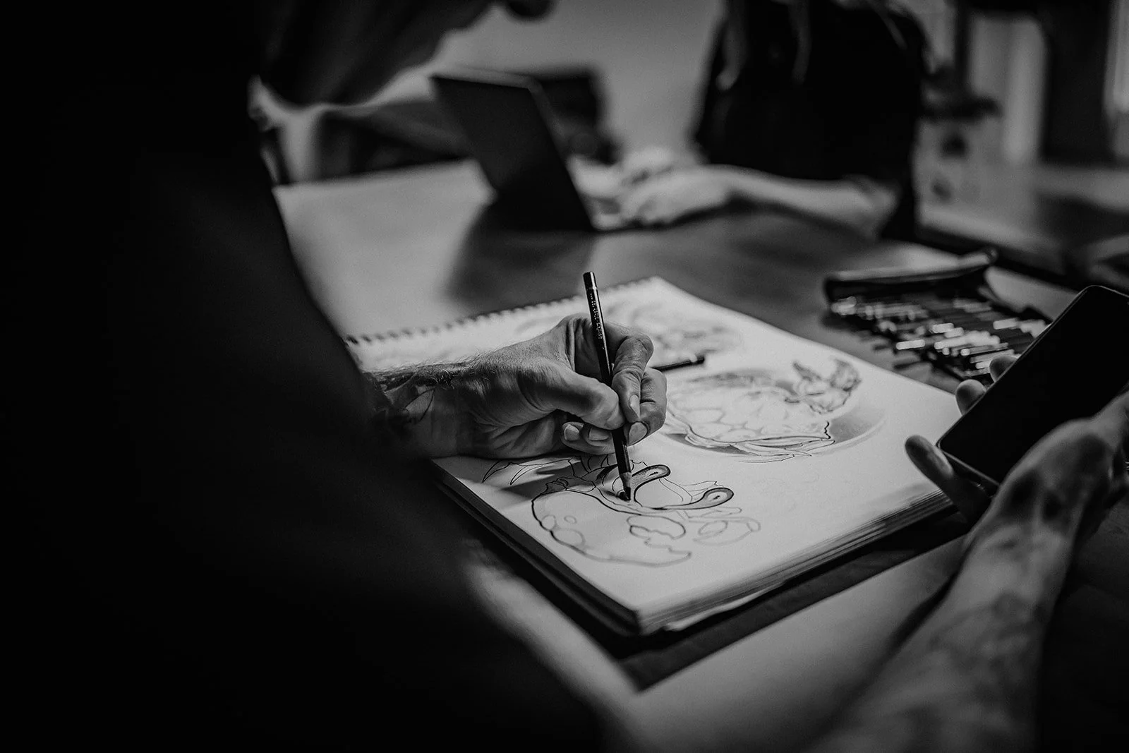 Black and white photo of an artist drawing a colorful sketch in a sketchbook, with various art supplies and devices on a table.