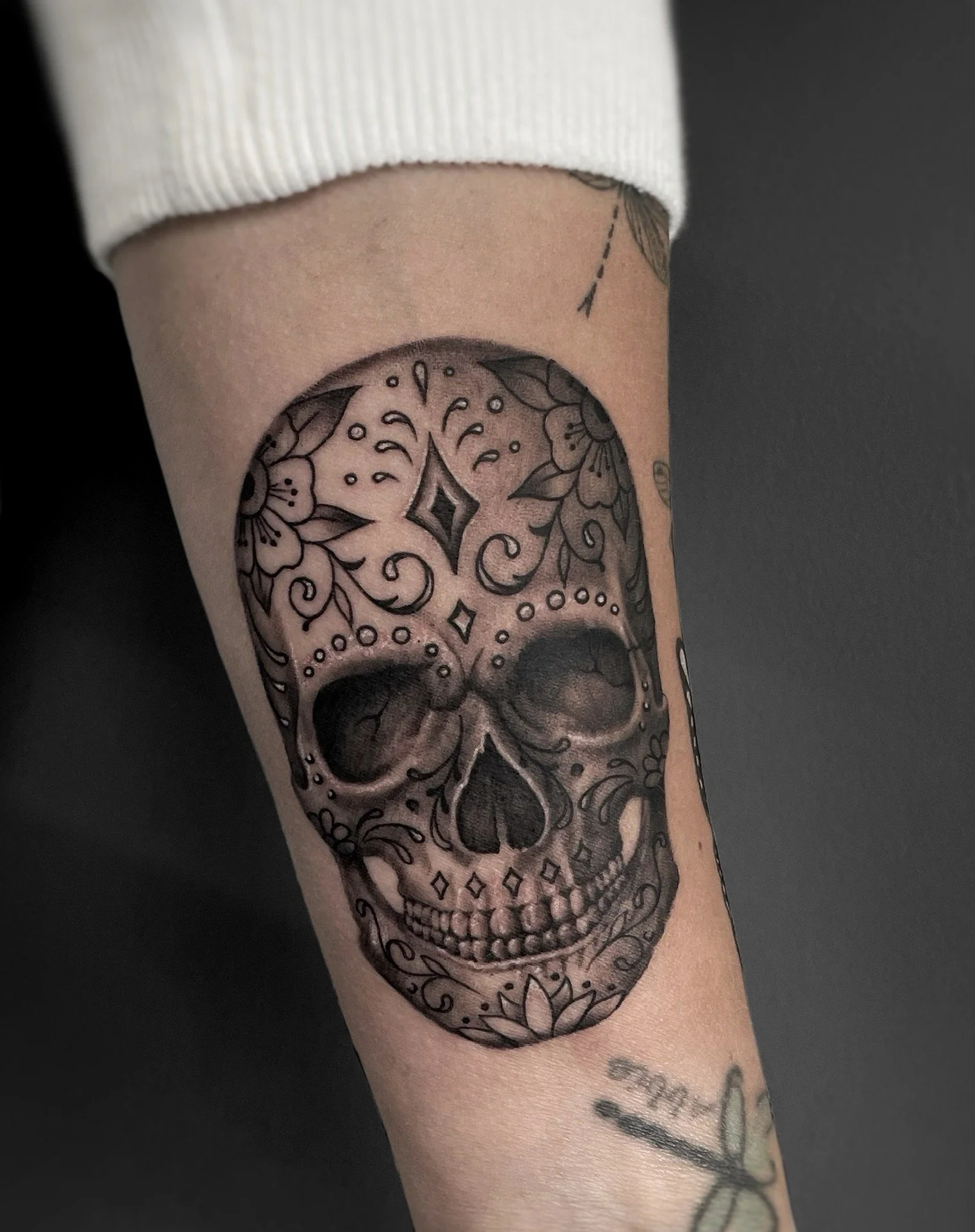 Black and gray tattoo of a sugar skull with floral and decorative patterns on a person's forearm.