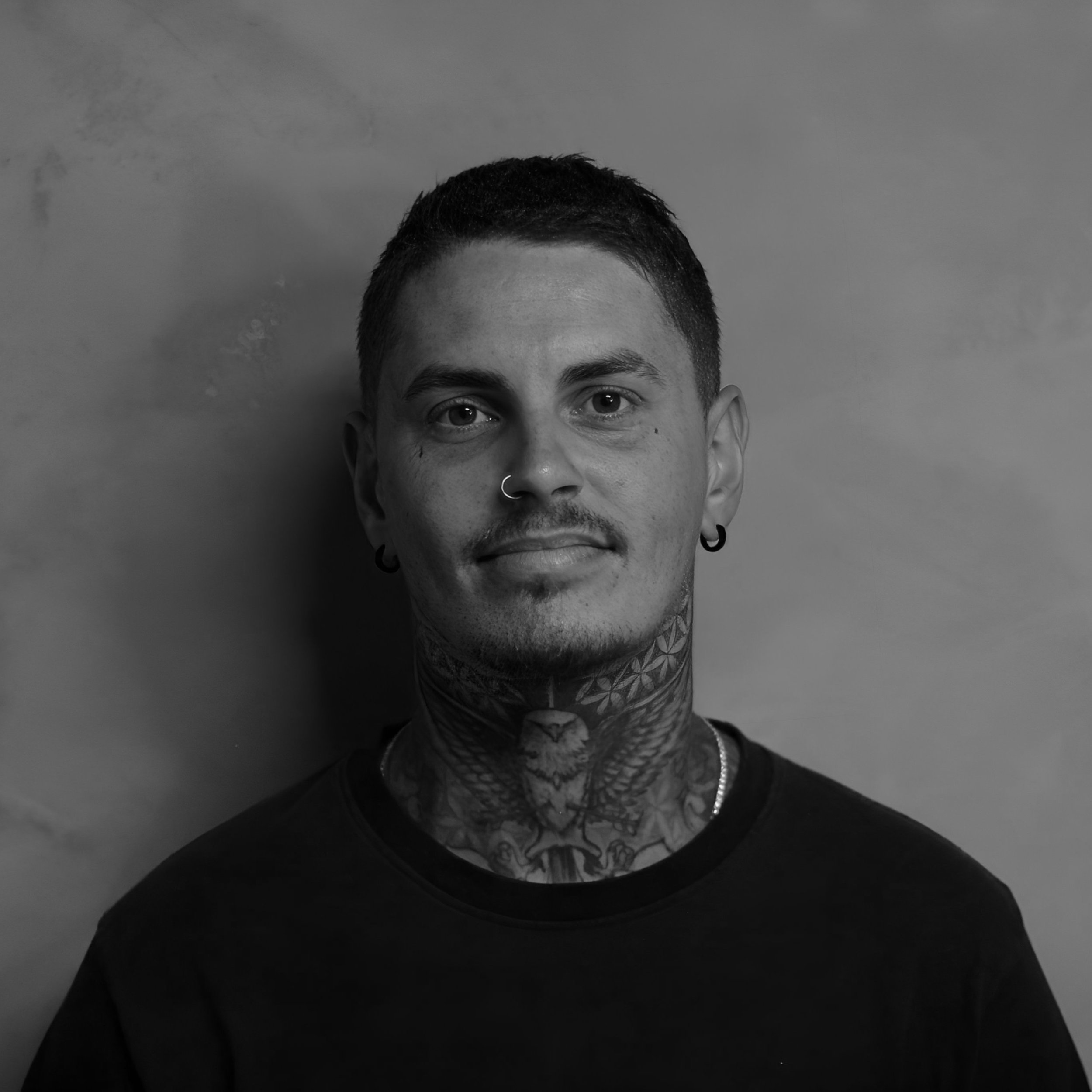 Black and white portrait of a young man with tattoos on his neck, wearing earrings, a nose ring, and a black shirt, standing against a plain wall.