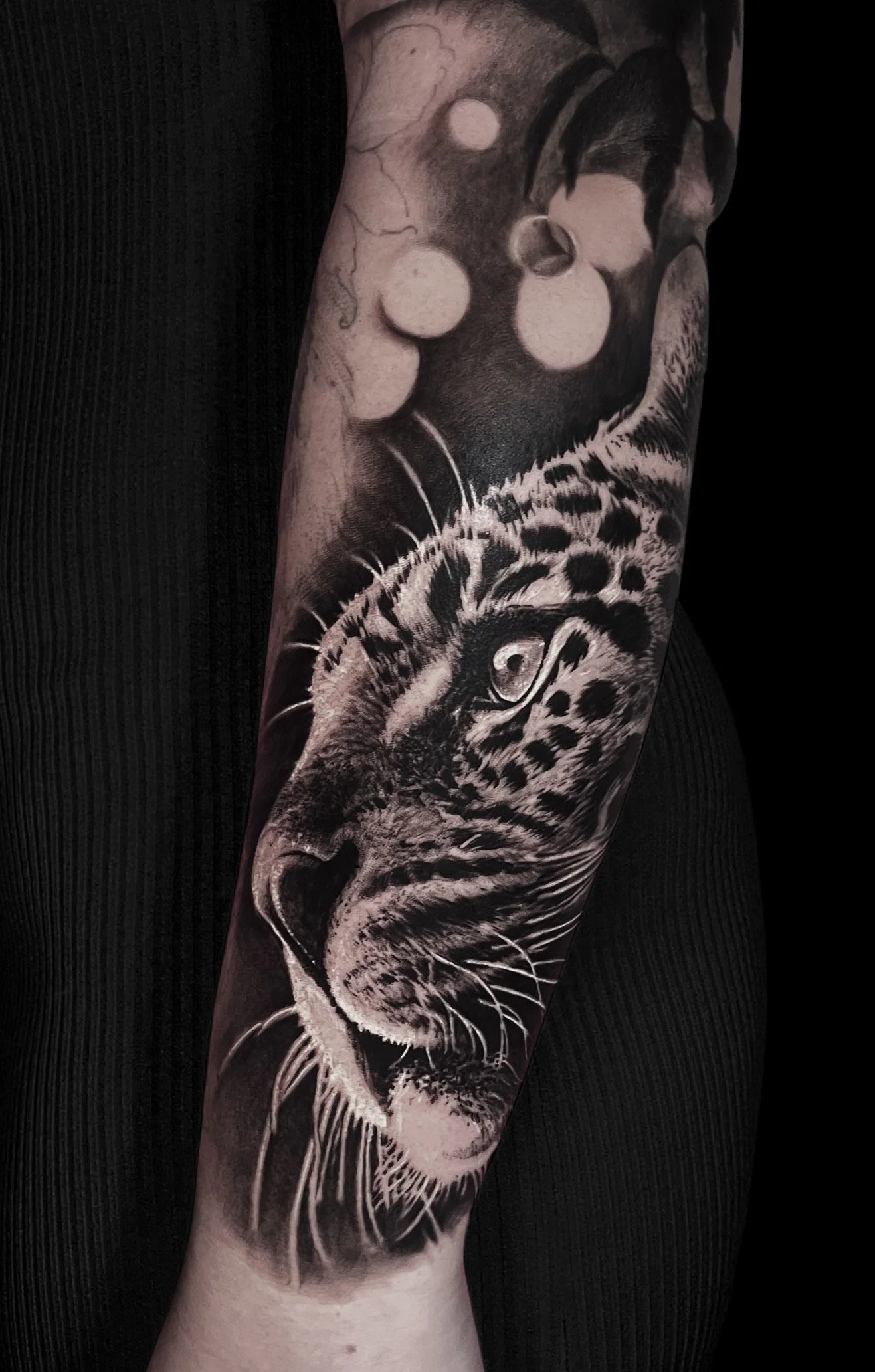 Black and white tattoo of a leopard's face on a person's arm, with detailed fur, whiskers, and intense eyes.