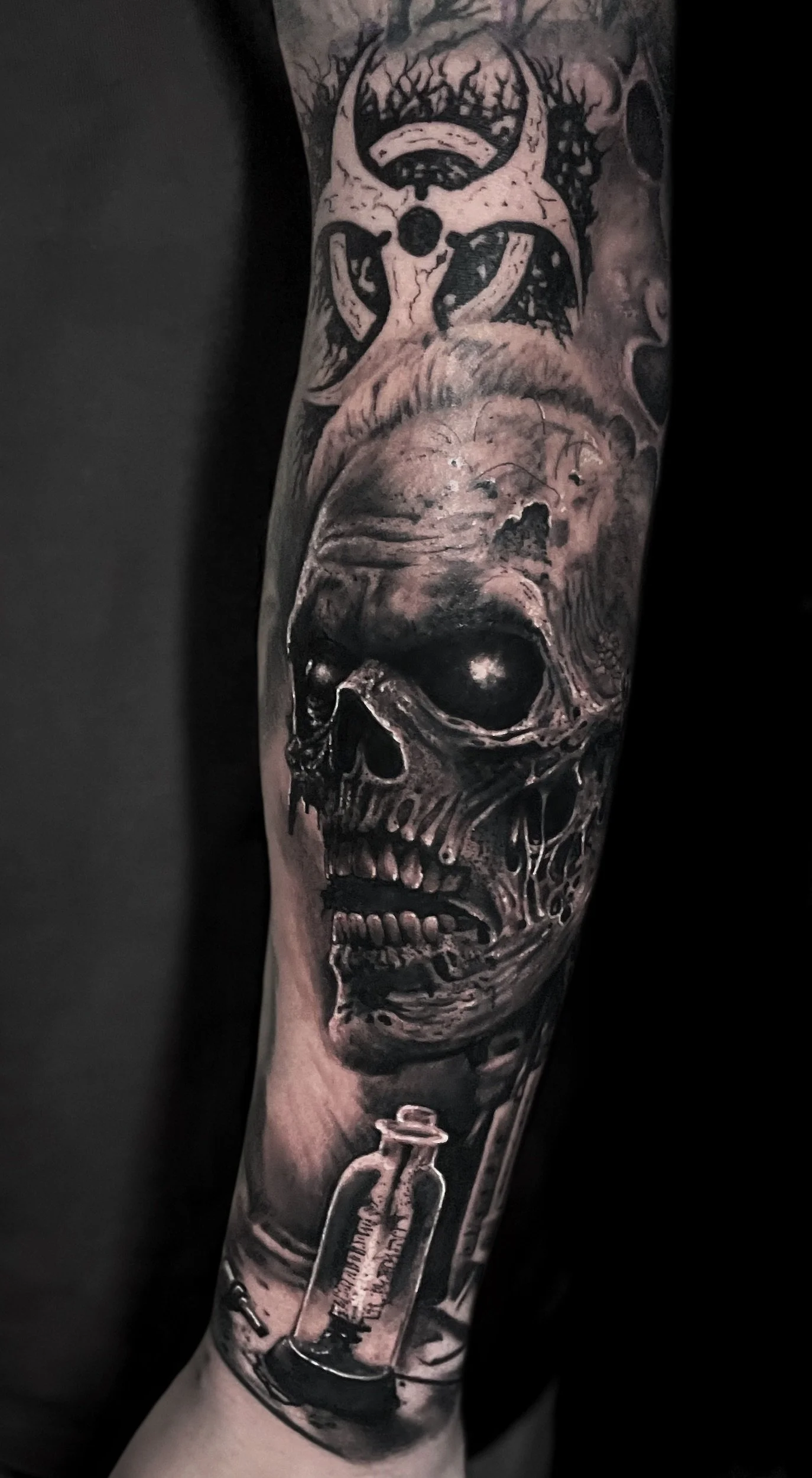 A detailed black and gray tattoo on a person's arm featuring a human skull with a syringe below it and a head with a mustache and a biohazard symbol above it.