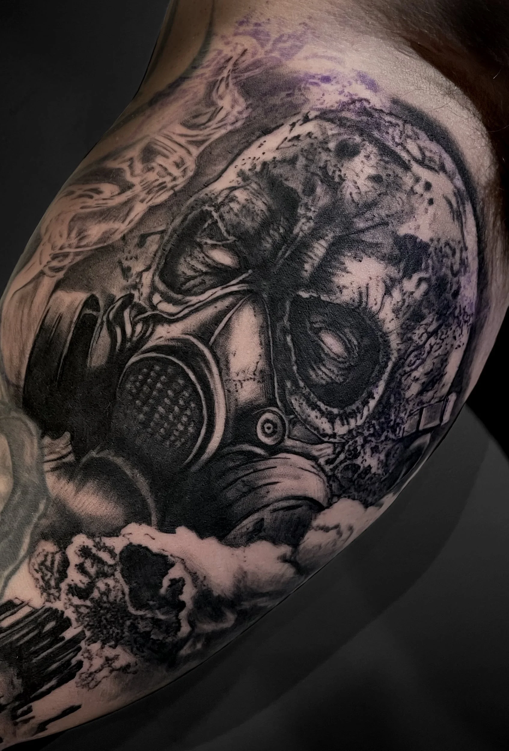 Tattoo of a skull wearing a gas mask with clouds and abstract elements in black and gray ink.