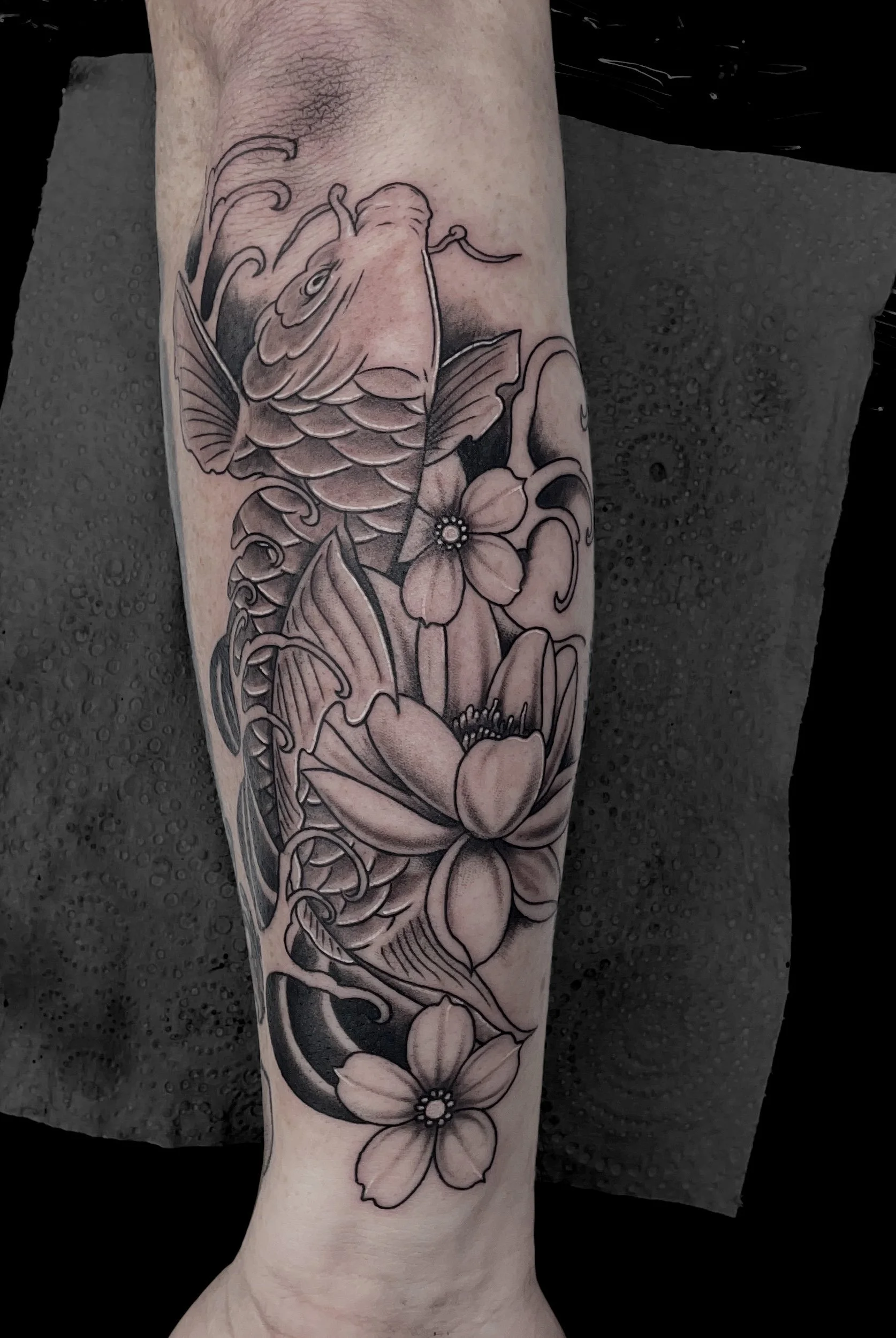 Black and gray tattoo on forearm featuring a koi fish and several large flowers with leaves.