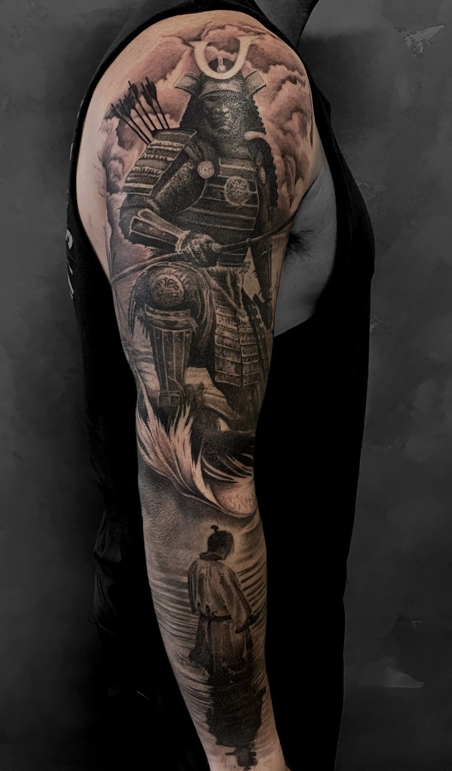 A detailed black and gray tattoo of a samurai warrior in traditional armor, holding a sword, with clouds and a warrior in traditional attire standing by a body of water in the background.