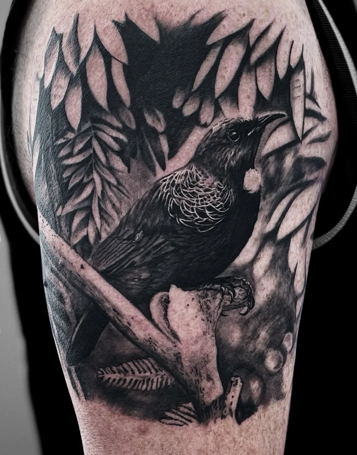 Black and gray tattoo of a bird with a scoop of food in its beak, perched on a branch amidst leaves.