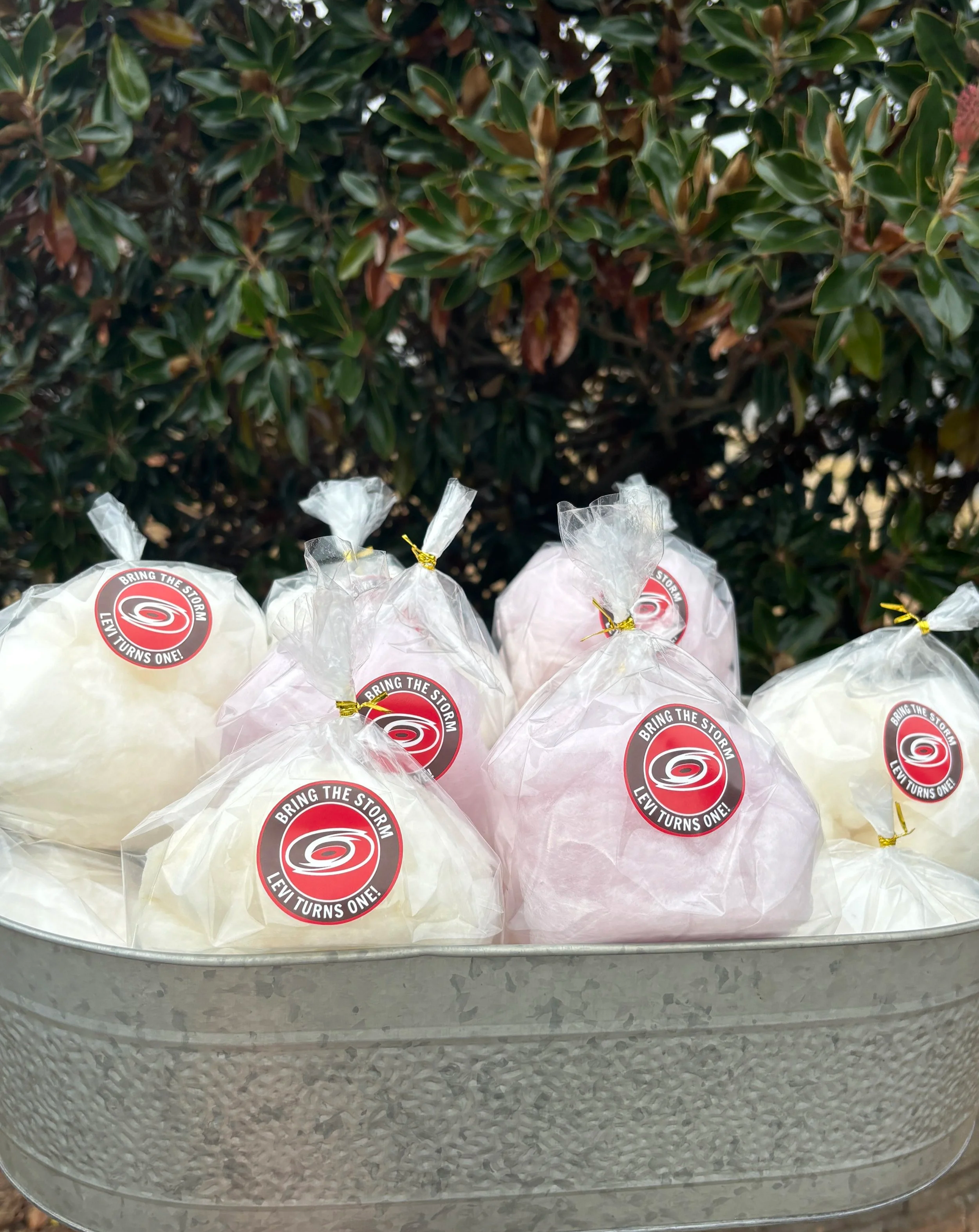 A hand holding a gray metal tray with individual wrapped pieces of cotton candy, each with a red and black sticker that reads "Bring the Storm, Levi Turns One!" in front of leafy bushes and dry grass.