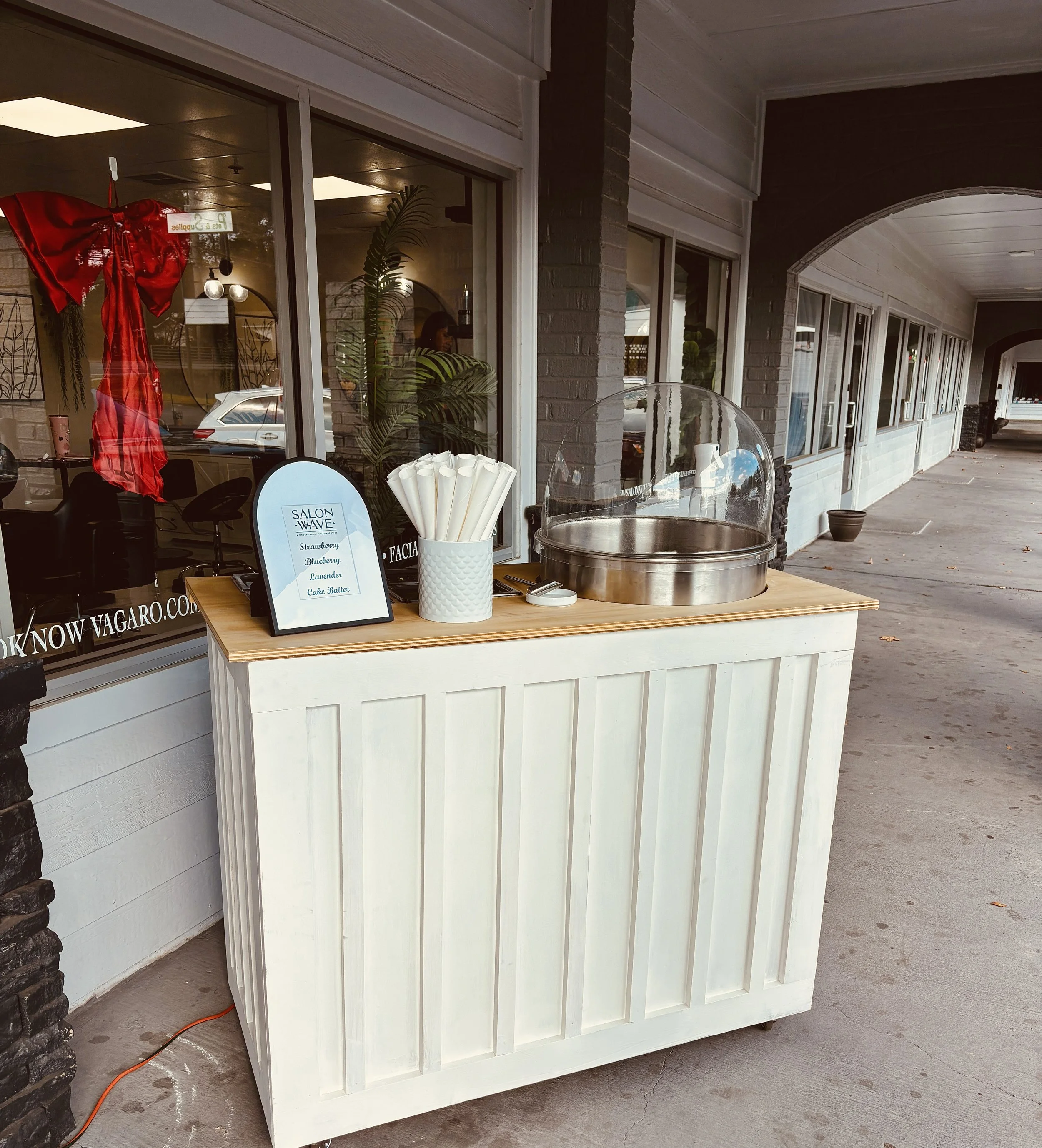 Outdoor dessert station with a cotton candy machine, a container holding white paper cones, and a sign advertising flavors like strawberry, blueberry, lavender, and cake batter for a salon wave event.