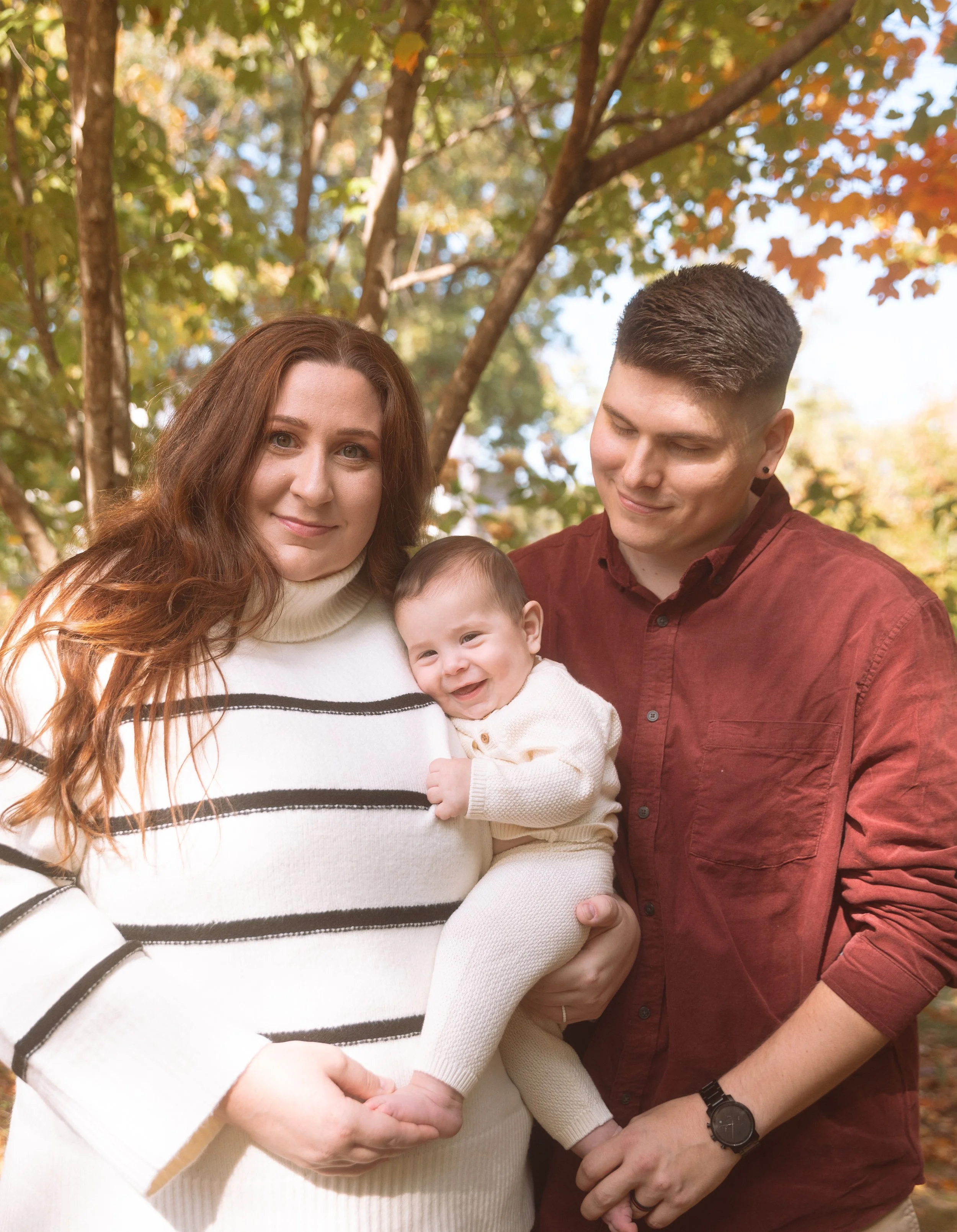 A family of three outdoors during fall; a woman with long red hair holding a smiling baby, a man with short dark hair looking at the baby, surrounded by autumn trees.