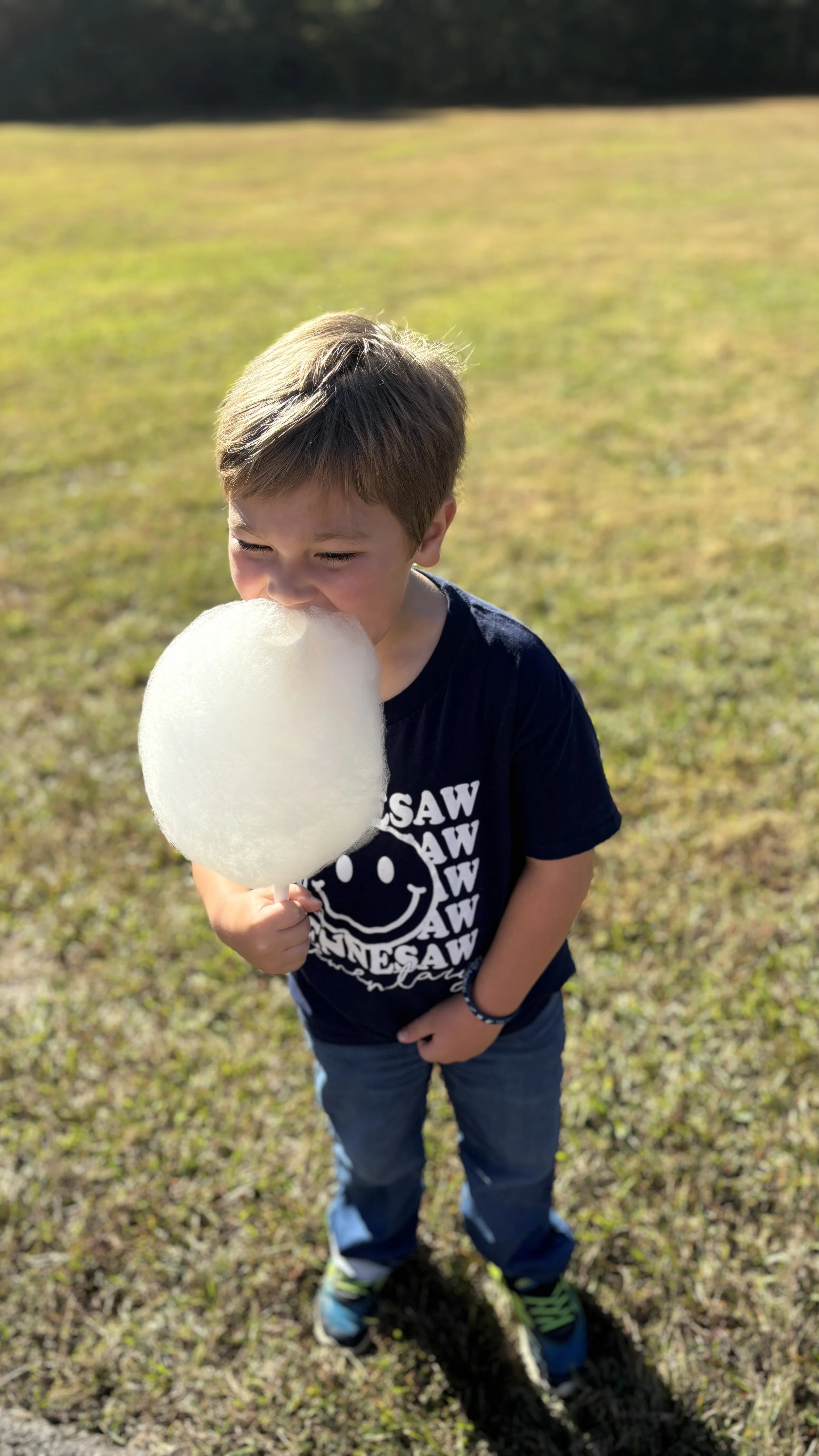 A young boy with brown hair holding a large stick of white cotton candy near his mouth while standing on a grassy field, smiling and enjoying a sunny day.