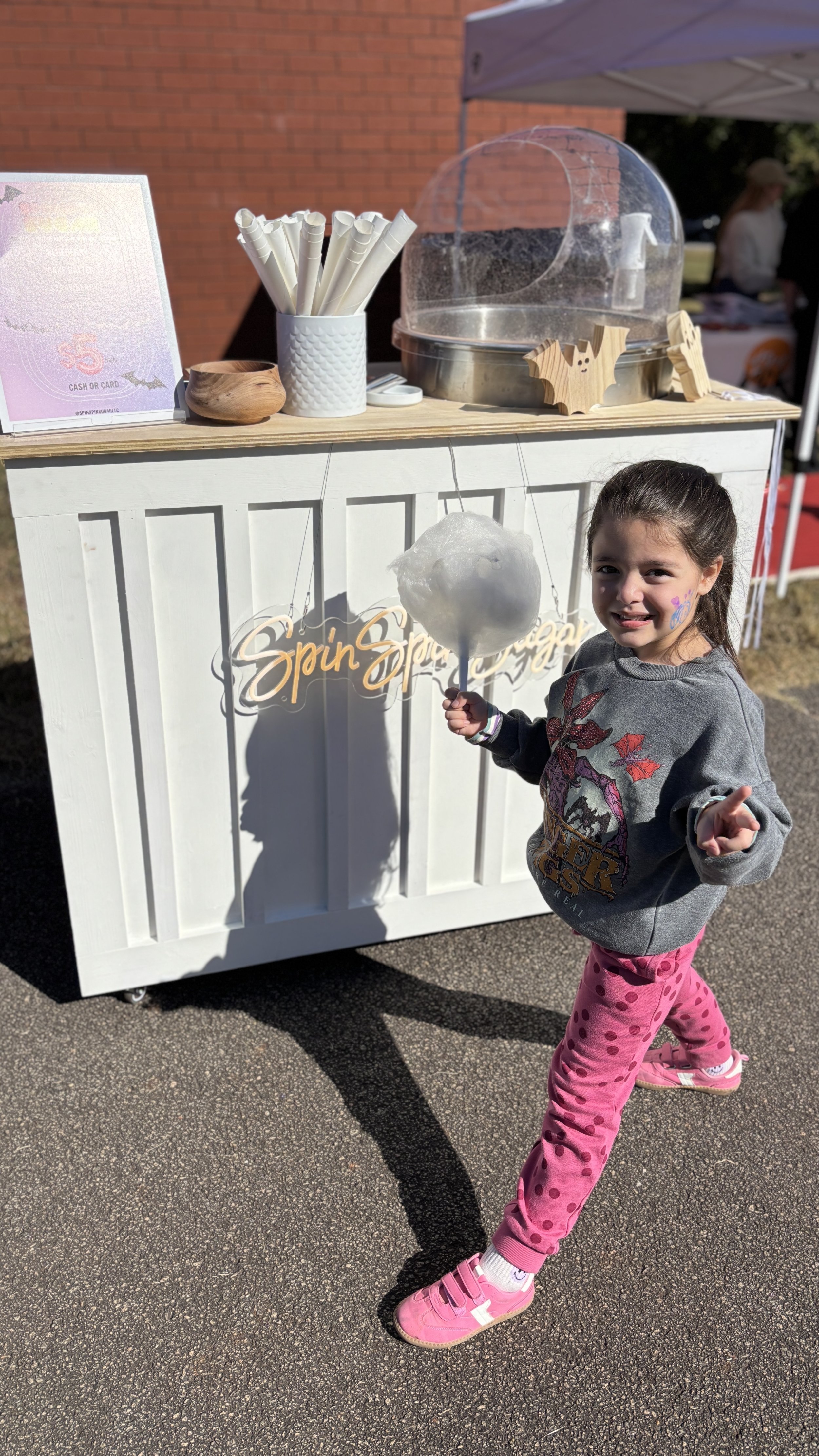 A young girl standing next to a cotton candy stand, holding a large piece of cotton candy on a stick. She is smiling, with face paint on her cheek, wearing a gray sweatshirt and pink polka dot leggings. The stand is decorated with a sign that reads '