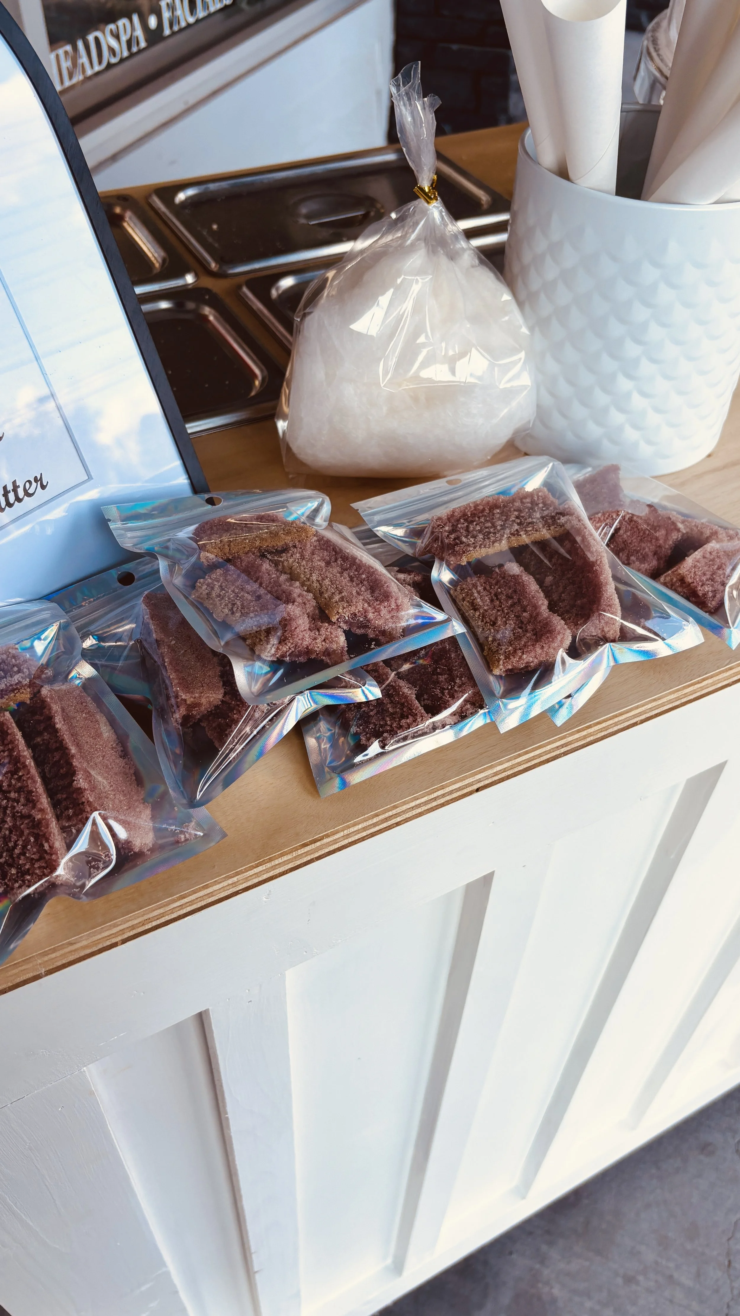 Clear plastic bags filled with salted licorice candies on a wooden counter at a candy shop, with a bag of white sugar and a white container with rolled paper nearby.