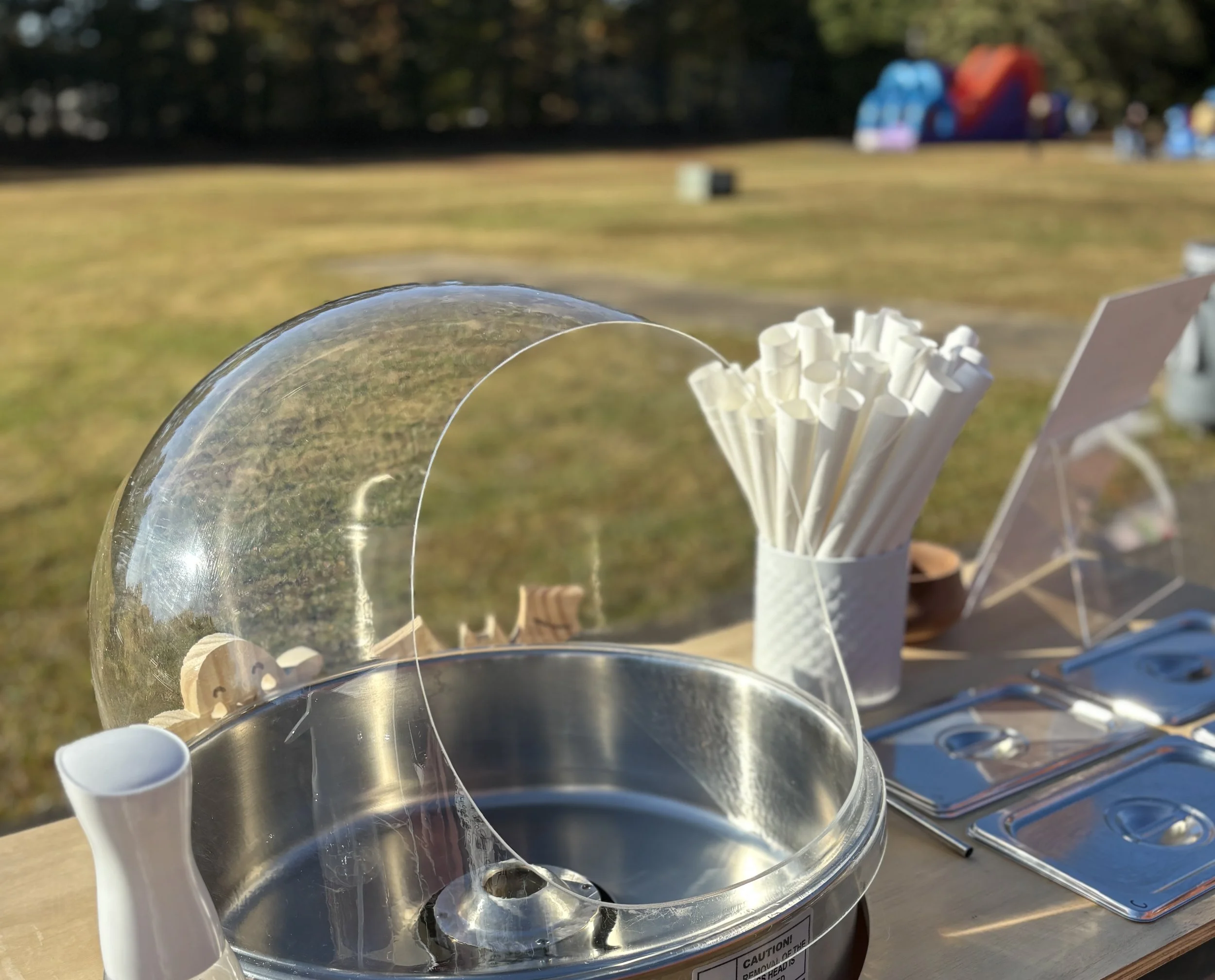 Cotton candy machine with a clear cover, placed on a wooden table outdoors. Behind it are white paper straws in a white container, some stainless steel containers, and a small brown bowl. The background shows a grassy field with trees and a colorful 