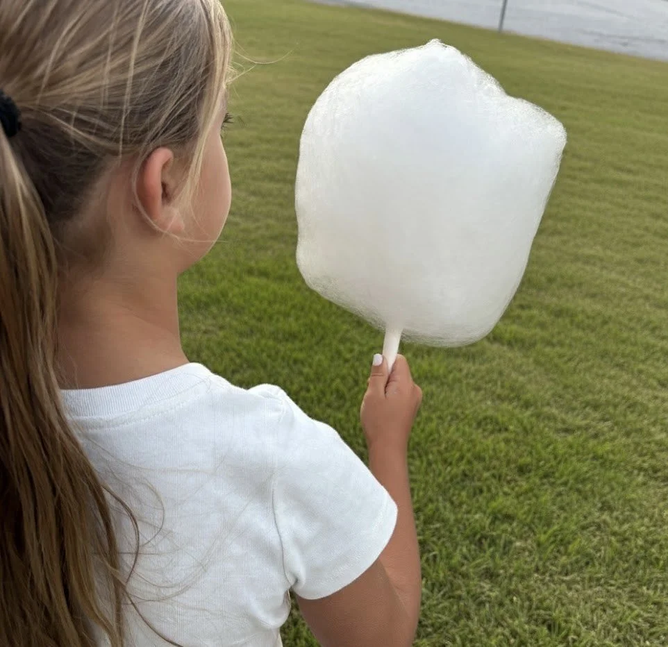 A girl with long blonde hair in a ponytail holding a large piece of cotton candy on a stick, standing outdoors on a grassy area near a body of water.