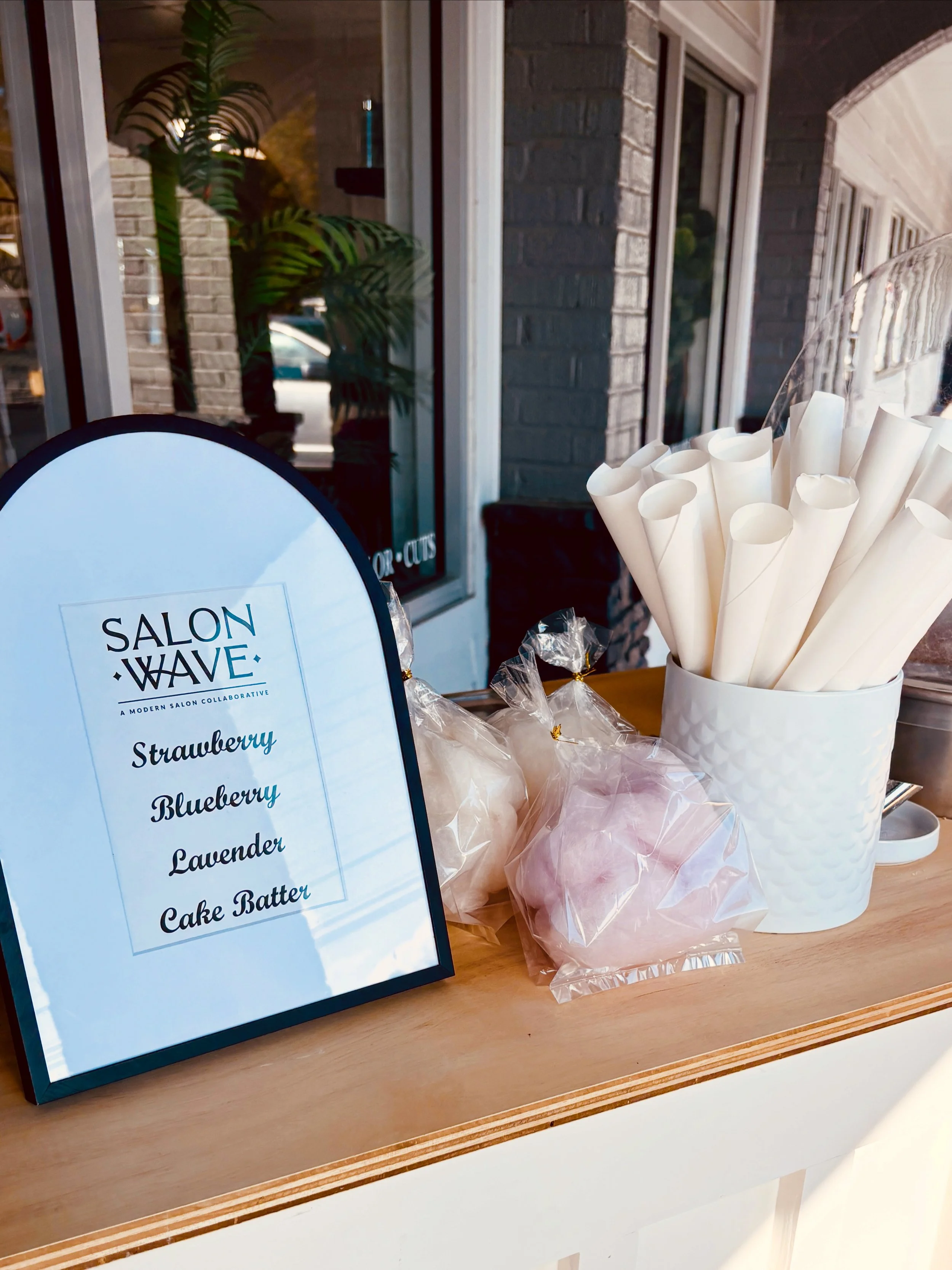 A tabletop display at a bakery featuring a sign for Salon Wave with flavors including strawberry, blueberry, lavender, and cake batter. There are pink and white soap or bath bombs wrapped in plastic and tied with gold, and a white container holding r